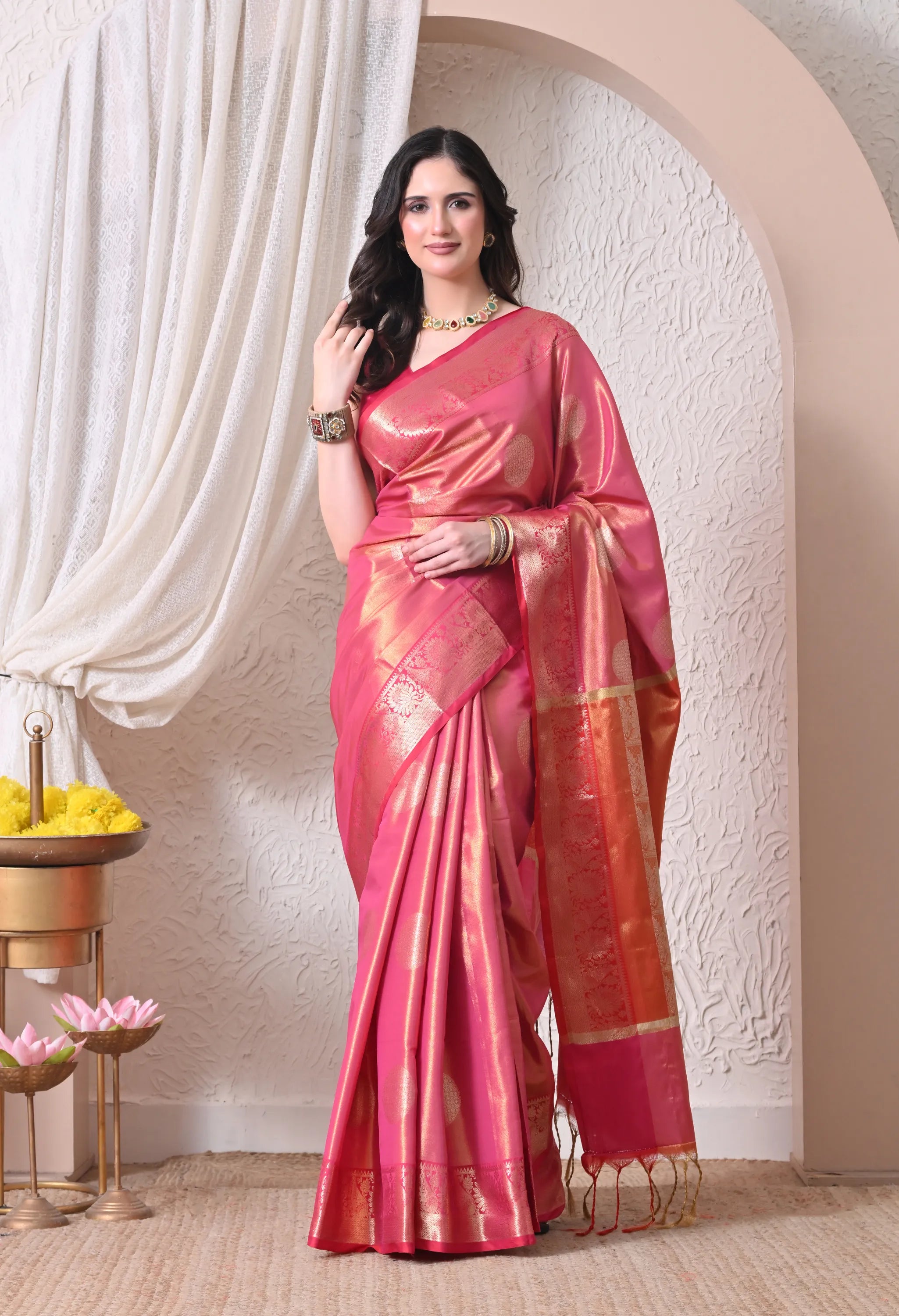 A woman posing in a vibrant pink silk blend saree featuring large circular gold zari motifs, a broad intricately woven gold border, and a matching tassel-trimmed pallu.
