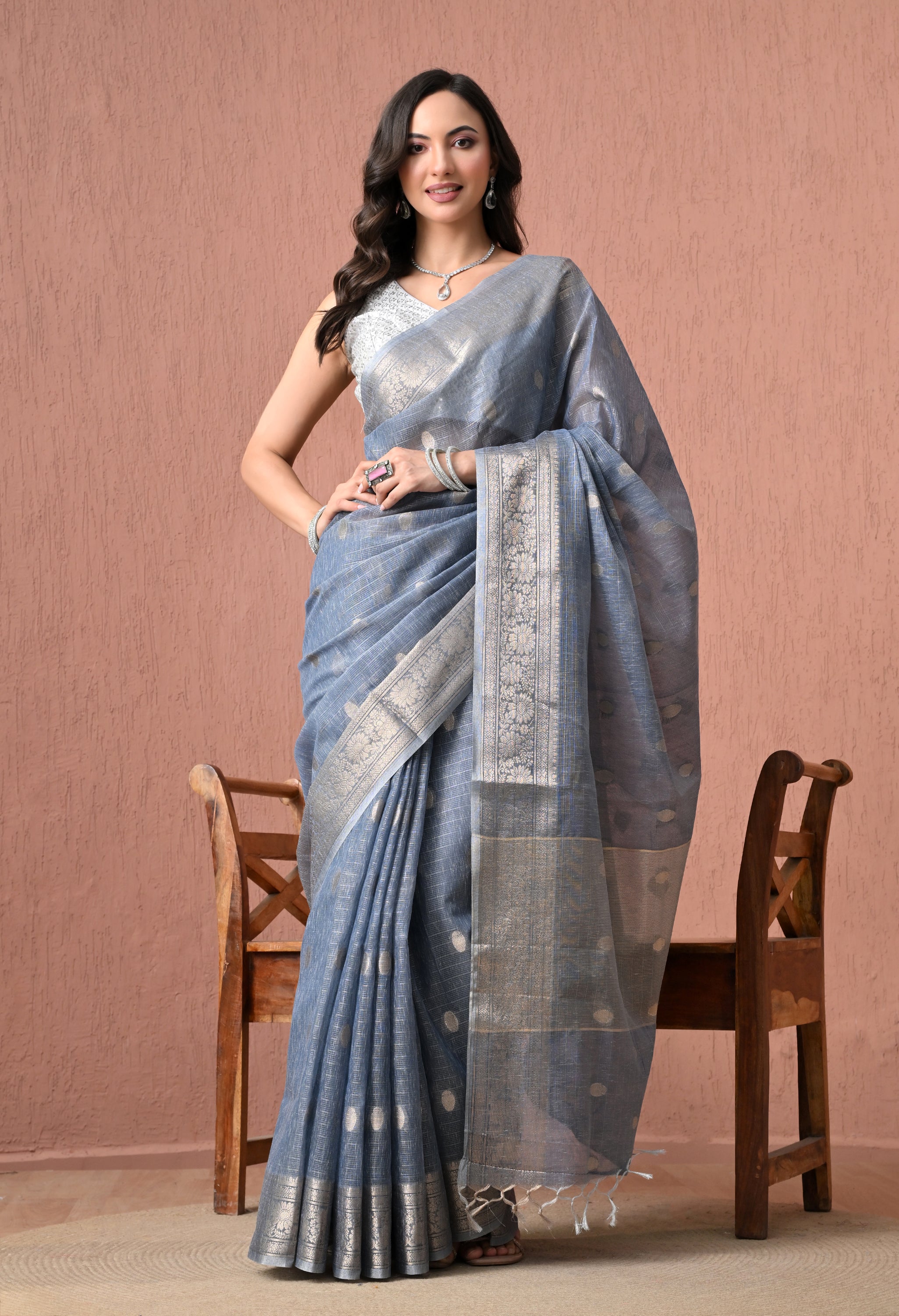 A woman modeling a steel blue cotton saree with an intricate silver zari border, standing in front of a textured pink background.
