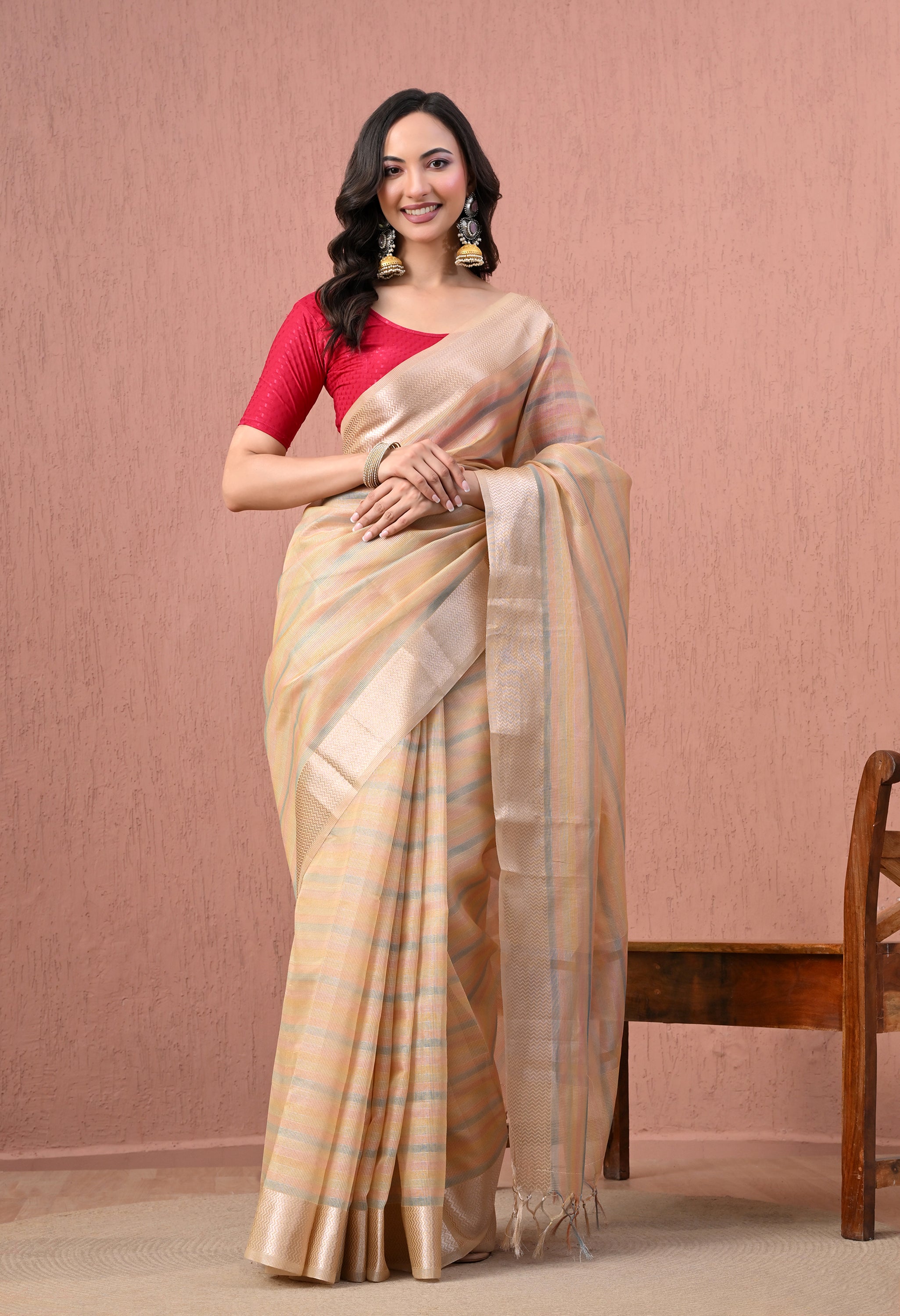 A full-length studio photograph of a smiling woman posing in a light beige pure cotton saree featuring pastel multicolor stripes and a wide metallic gold zari border. She is wearing a contrasting bright fuchsia pink half-sleeved blouse, heavy traditional gold jhumka earrings, and a gold bangle, standing against a textured dusty pink backdrop next to a wooden chair.
