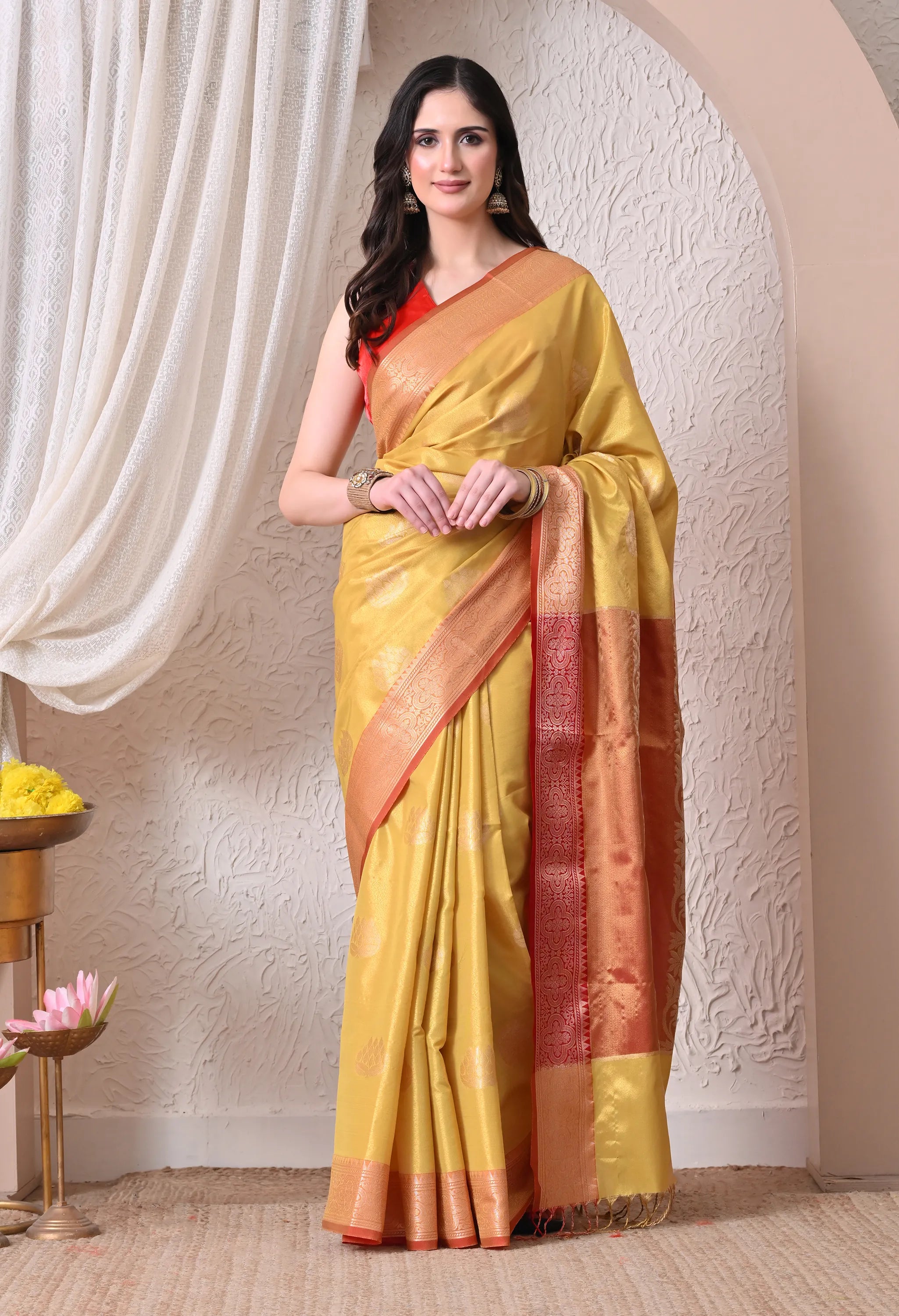 A woman posing in a vibrant mustard yellow silk blend saree adorned with golden lotus motifs and featuring a rich rust-red contrast border and tasseled pallu
