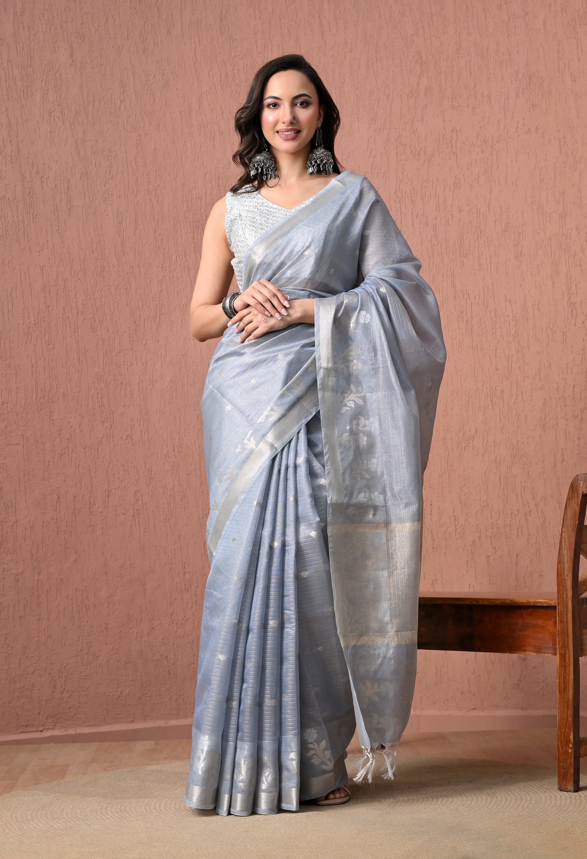 A full-length catalog photo of a model showcasing a powder blue pure cotton saree with woven silver zari floral patterns and a silver border. The saree has white tassel accents on the pallu. She is wearing a sleeveless white-and-silver blouse, large statement oxidised silver earrings, and dark bangles, posing against a textured dusty-rose background.
