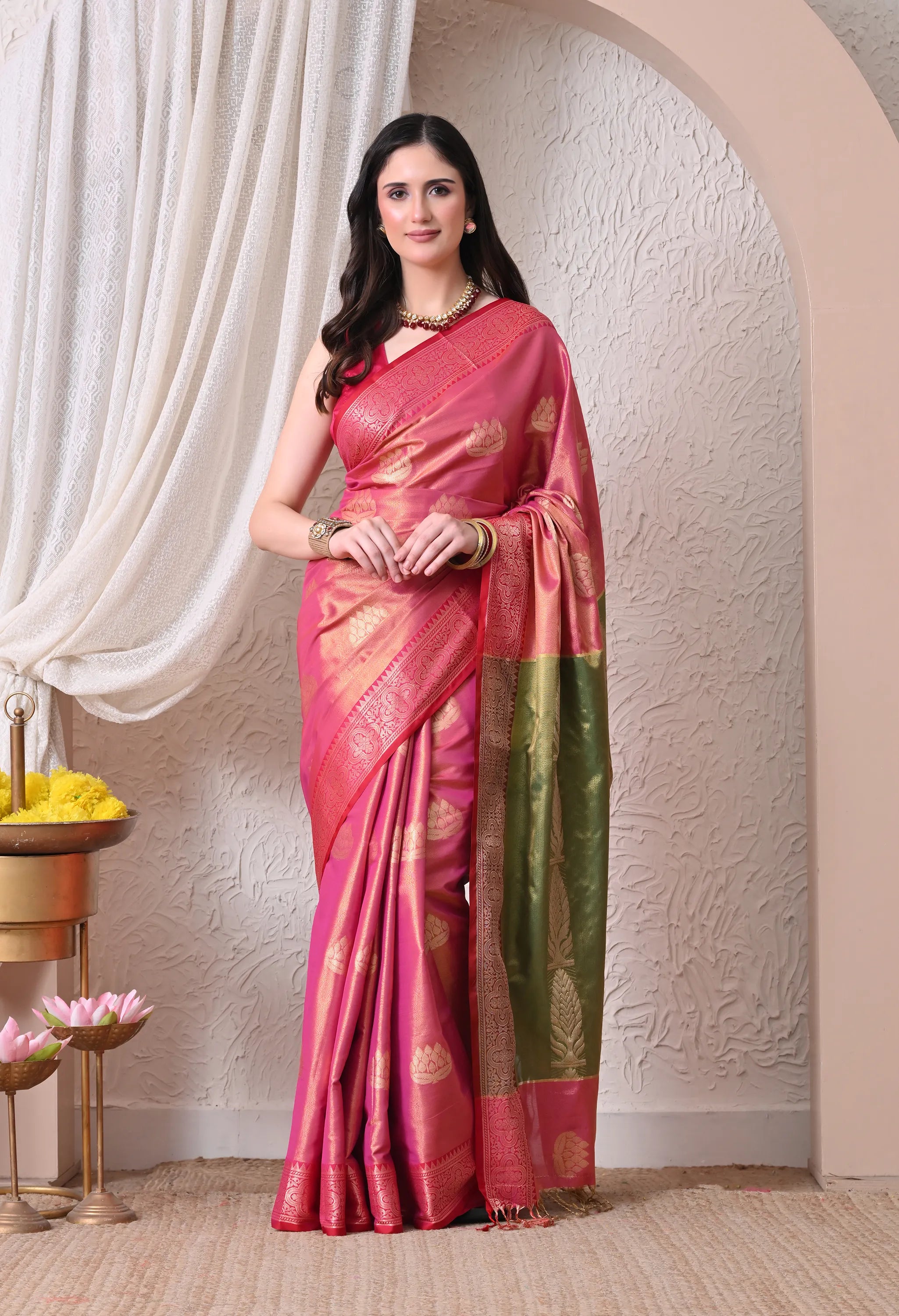 A woman posing elegantly in a Rani Pink silk blend saree with gold lotus motifs, featuring a striking contrast green and gold zari pallu.
