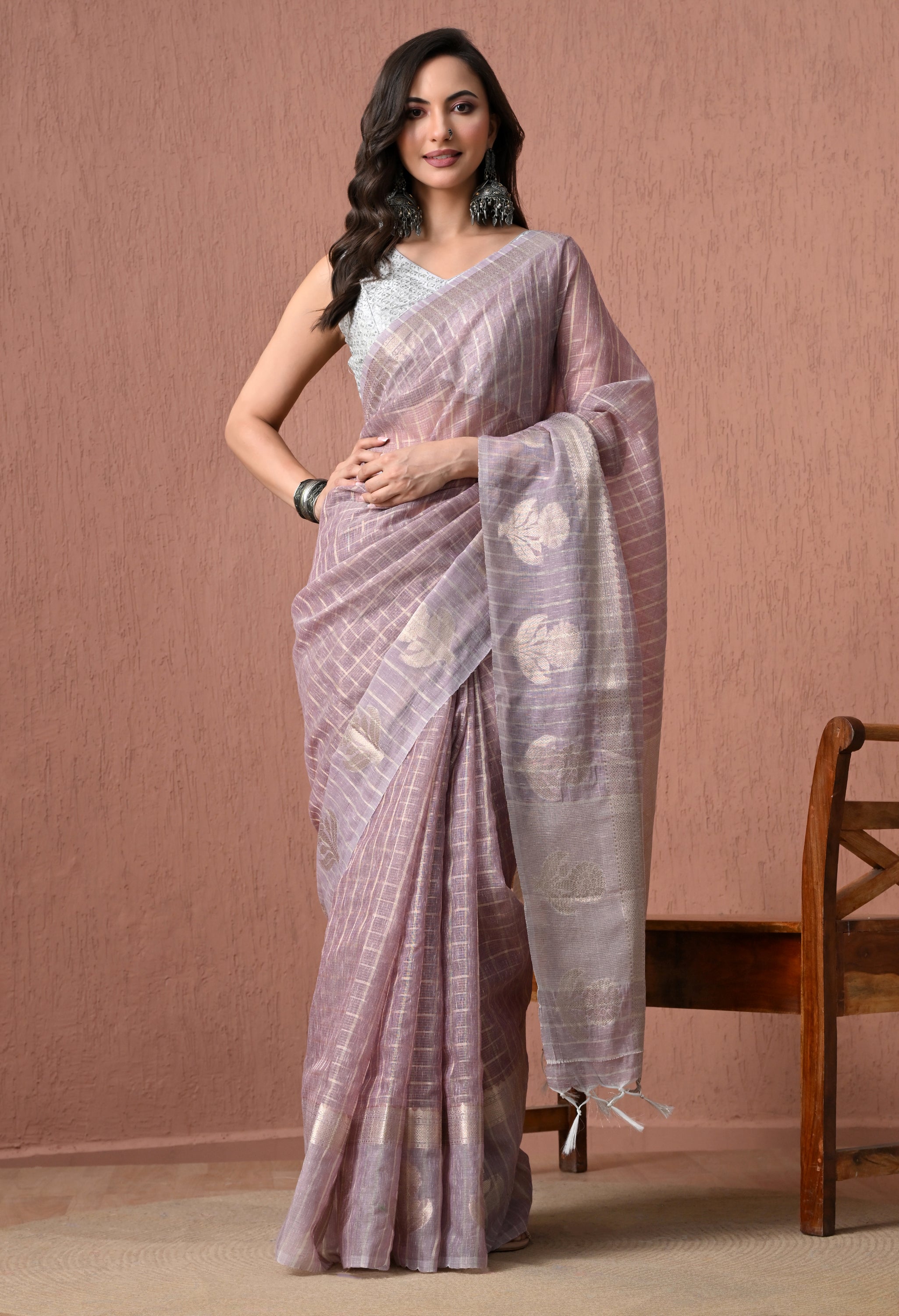 A full-length studio photograph of a woman posing in a mauve pure cotton handloom saree featuring fine metallic checks and a textured silver-grey pallu with woven floral motifs. She is wearing a white sleeveless blouse and large traditional silver earrings against a neutral peach backdrop.
