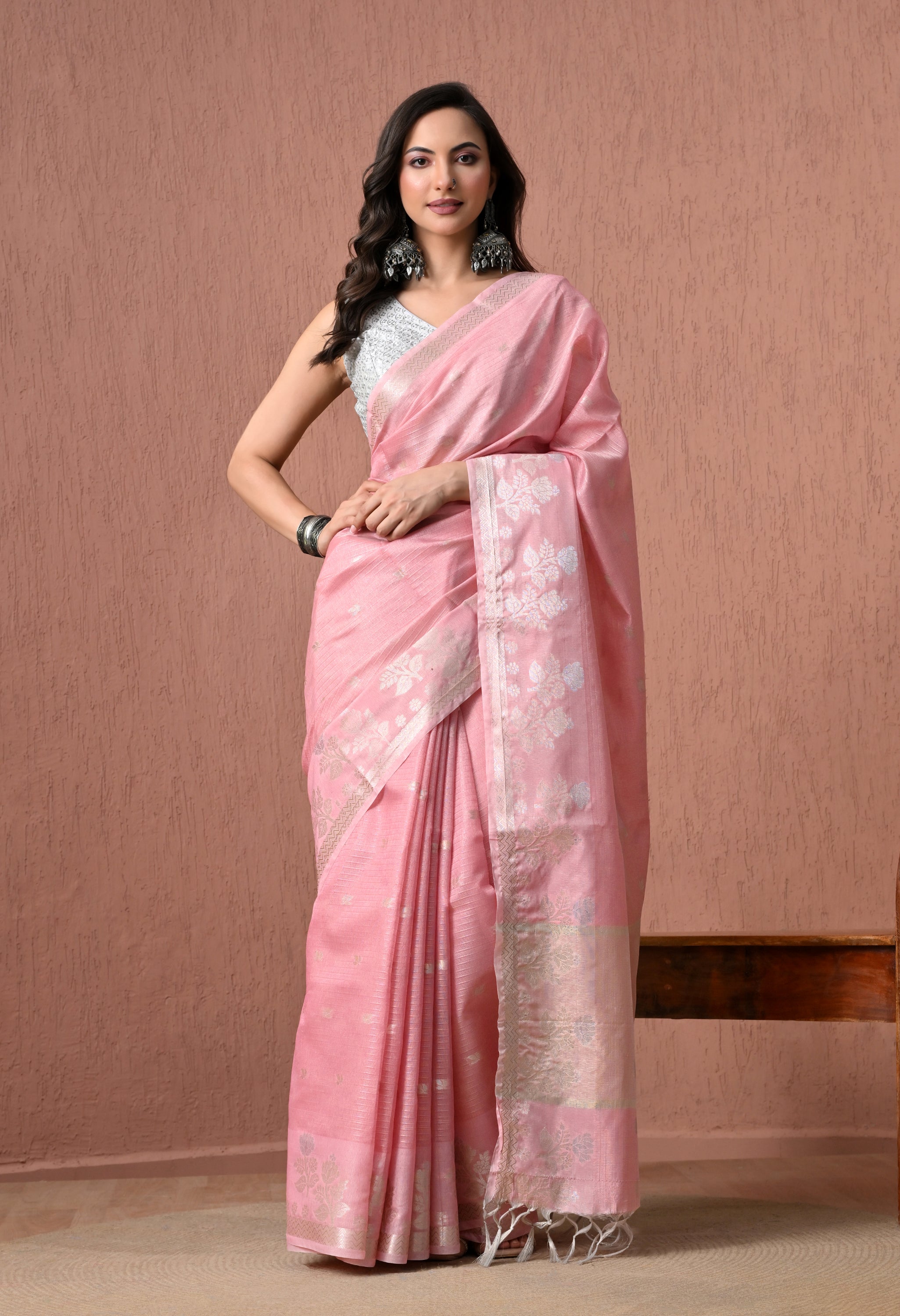 A full-length studio photograph of a woman modeling a bright blush pink pure cotton handloom saree. The saree features delicate silver zari floral motifs on the body and a heavily woven silver pallu with tassels. She pairs it with a sleeveless white textured blouse against a warm, textured background.
