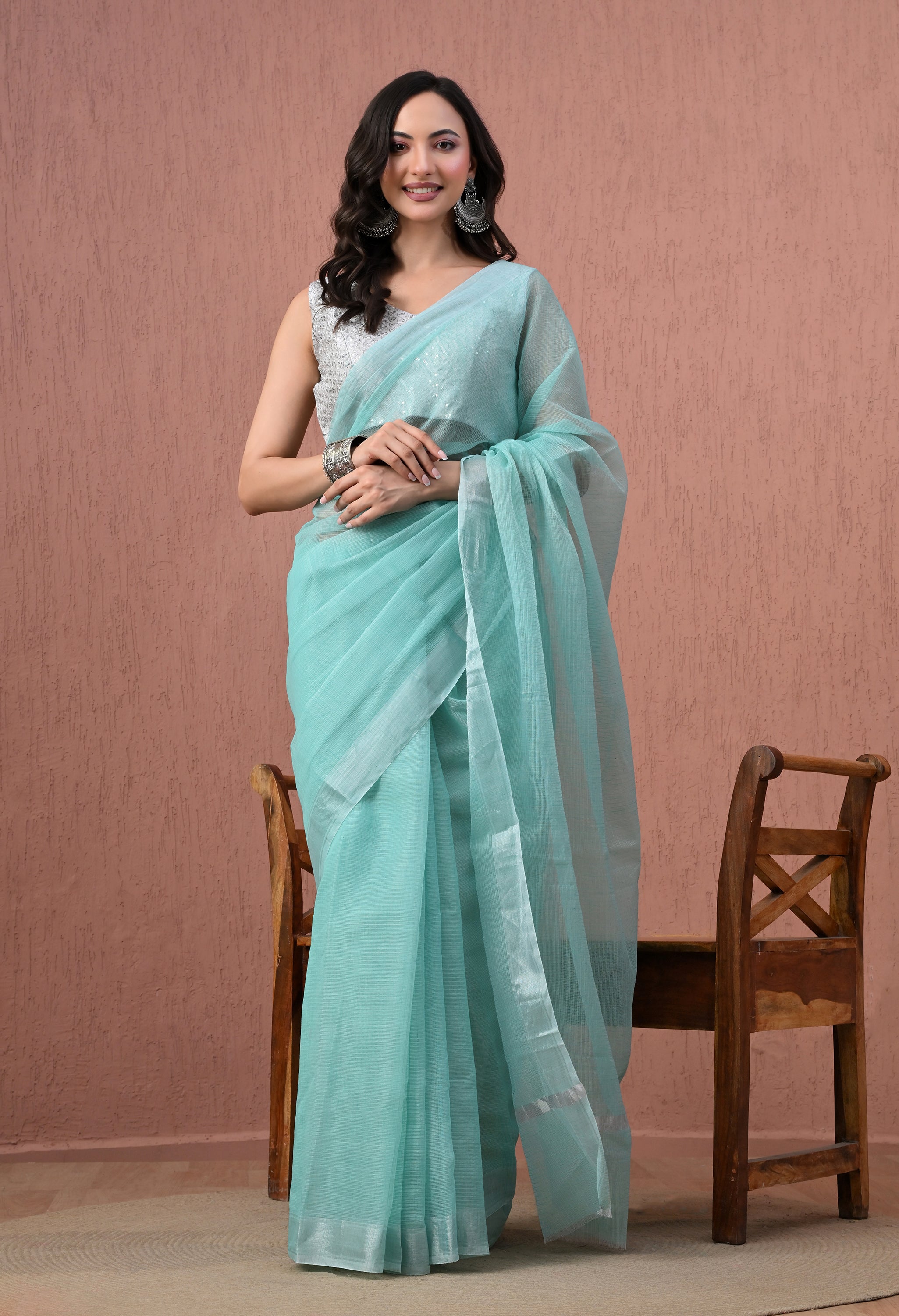 A full-length studio shot of a smiling model posing in a translucent, sea green pure cotton saree with a thin silver border. She is paired with a sleeveless silver sequined blouse, large oxidised silver earrings, and a matching chunky bracelet, standing in front of a textured dusty-rose background next to a wooden bench.
