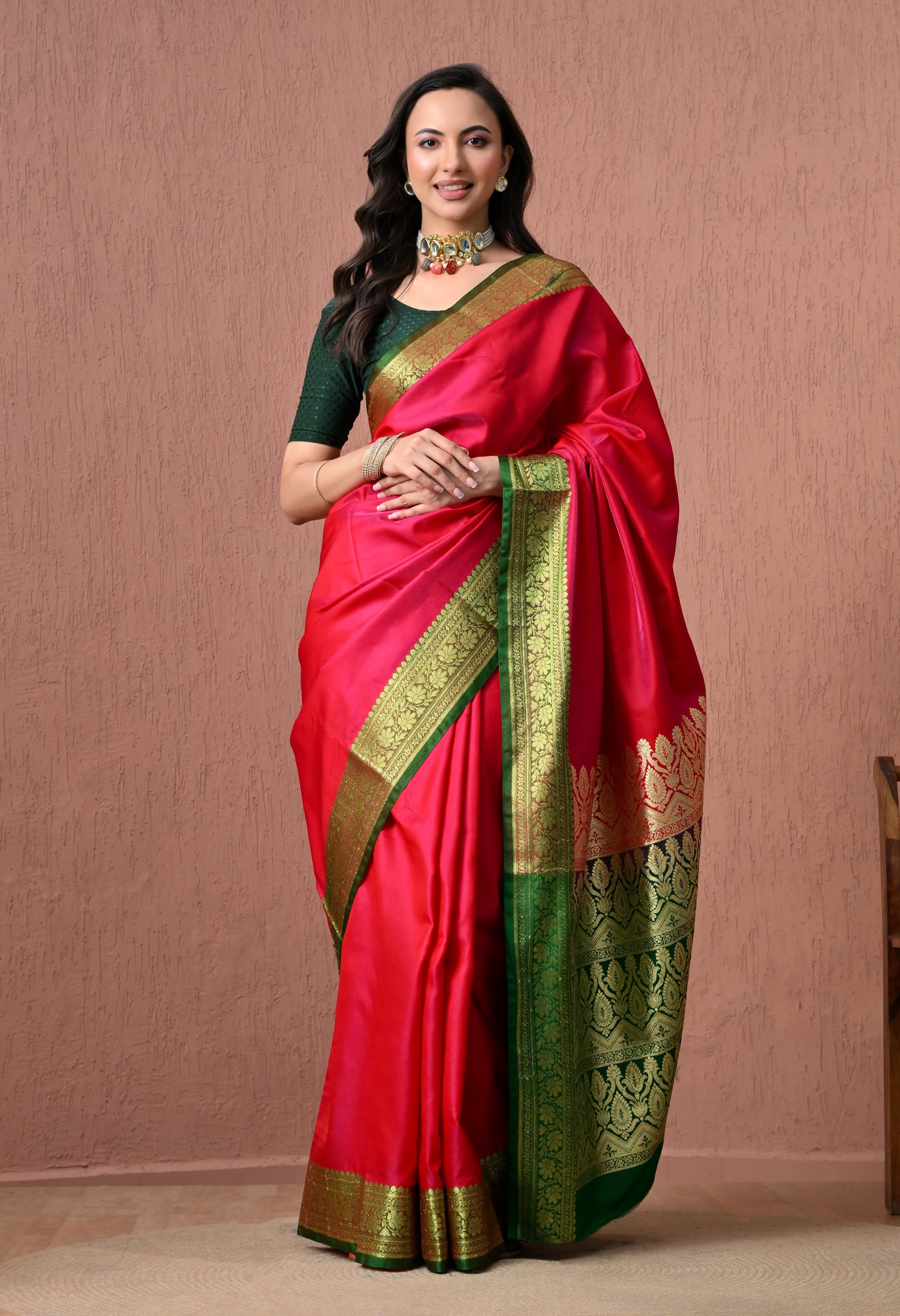 Full-length studio shot of a smiling model showcasing a vibrant rani pink pure silk Banarasi saree with a contrasting emerald green border and heavy gold zari pallu, paired with a green blouse and traditional jewelry.
