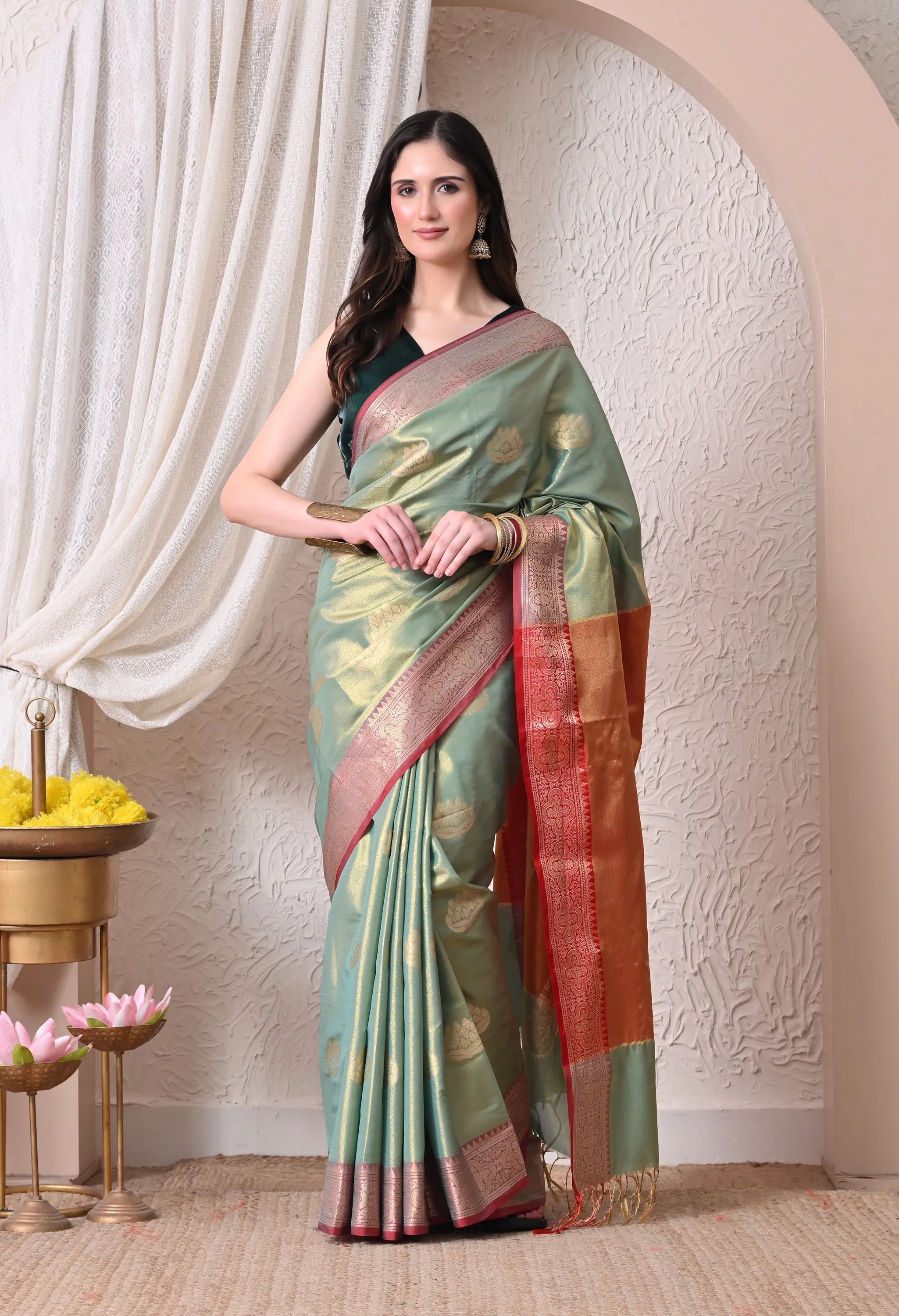 A woman posing in a sage green silk blend saree featuring golden lotus motifs, a rust-red contrast border, and a tasseled pallu
