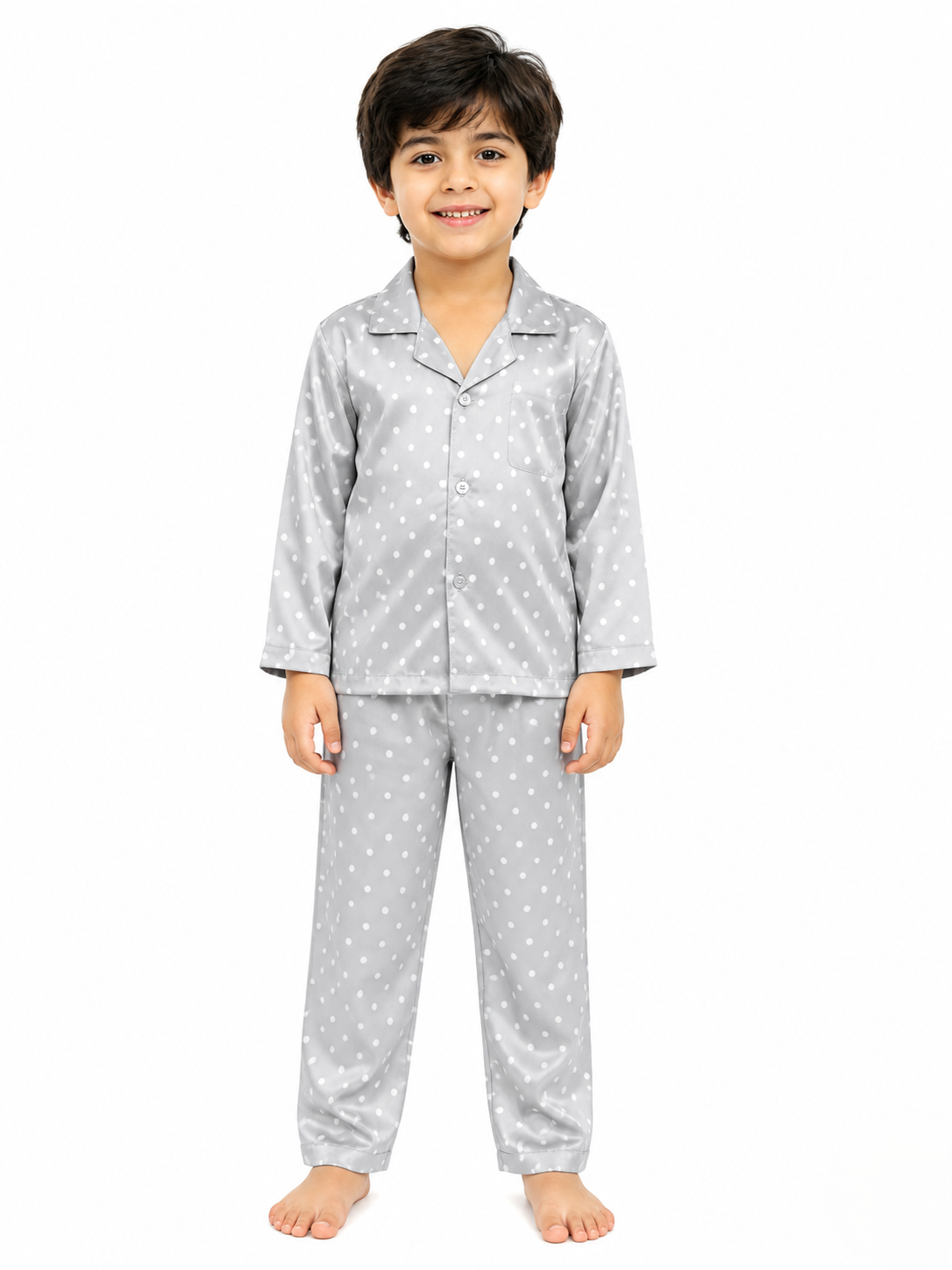 A young boy smiling and posing in a matching grey satin nightsuit set with a white polka dot print, featuring a long-sleeve button-down shirt with a notched collar and matching long pajamas against a solid white background.
