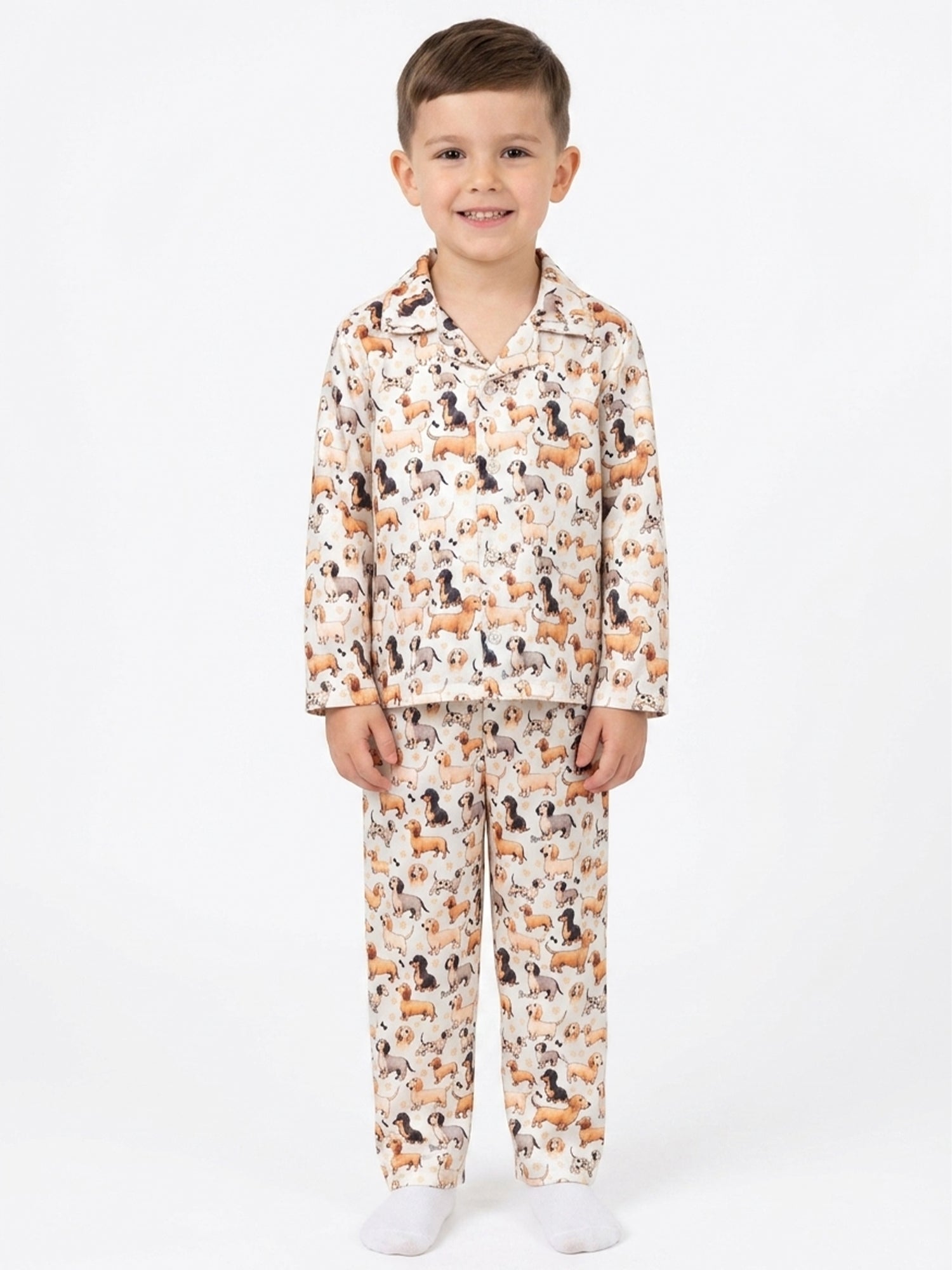 A young boy standing against a plain white background wearing a cream-colored satin pajama set with an all-over print of dachshund dogs.
