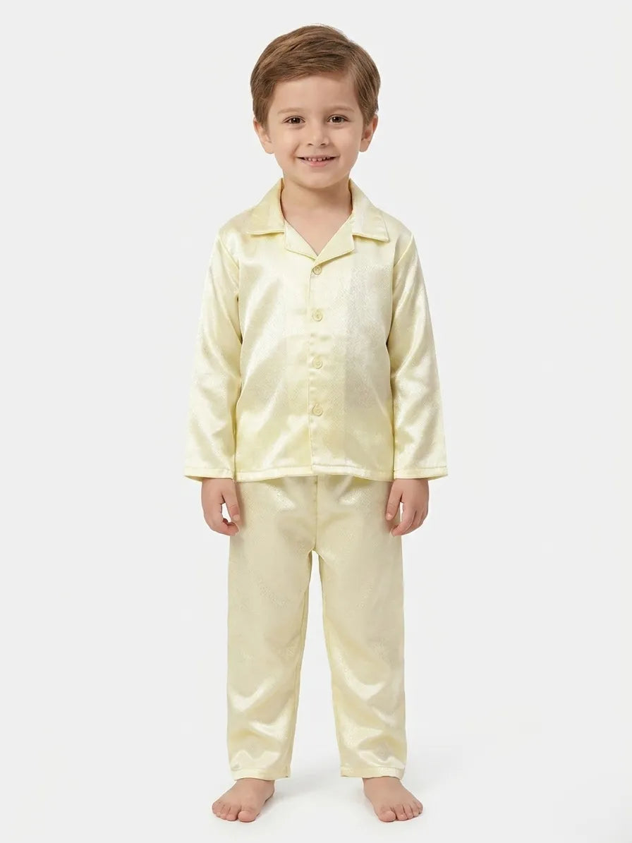A young boy standing against a white background wearing a light green, shiny satin night suit set consisting of a long-sleeved button-up shirt and matching pyjamas.
