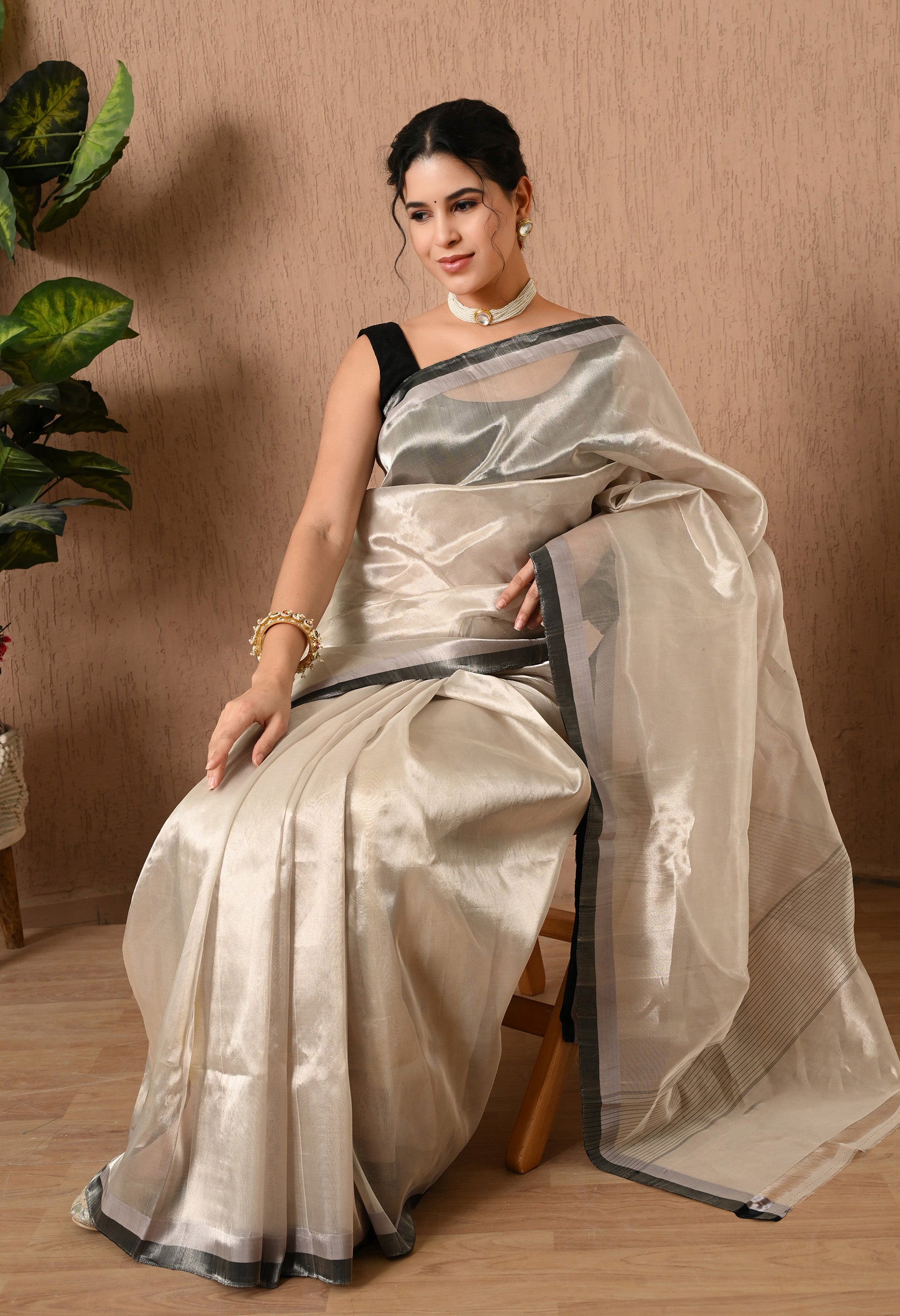 A woman sitting on a chair, modeling a shimmering silver tissue silk saree from The Weavers Shop by Raavi. The saree is made of a semi-transparent fabric with a brilliant metallic luster and a sleek black border. She is wearing a blouse, a choker, and a gold bangle, looking toward the camera with a smile against a neutral textured wall.