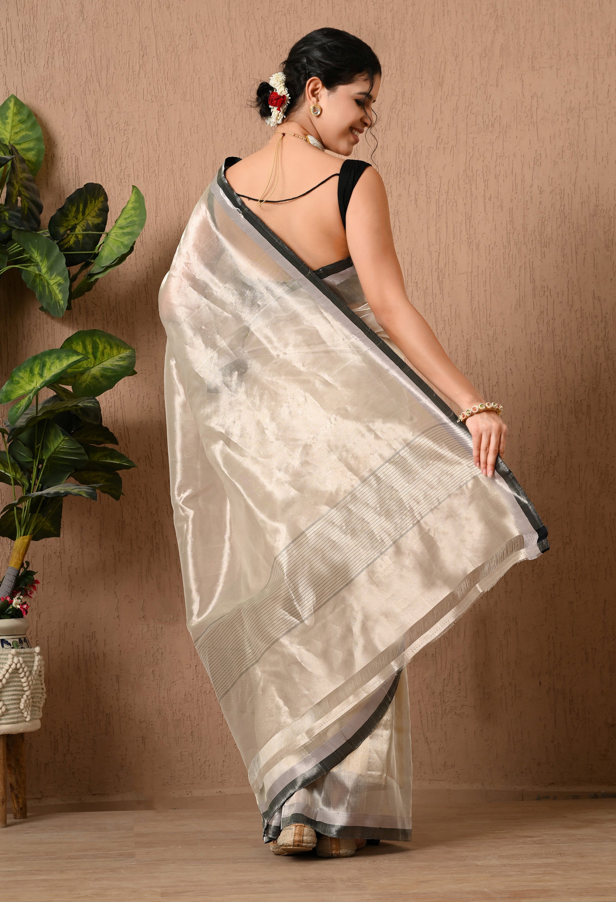 Back view of a woman showcasing a shimmering silver tissue silk saree from The Weavers Shop by Raavi. The saree is semi-transparent with a metallic luster and features a thin black and silver border along the hem and pallu. She wears a black blouse with a deep back and has her hair styled in a bun. The background consists of a textured tan wall and a potted green plant.