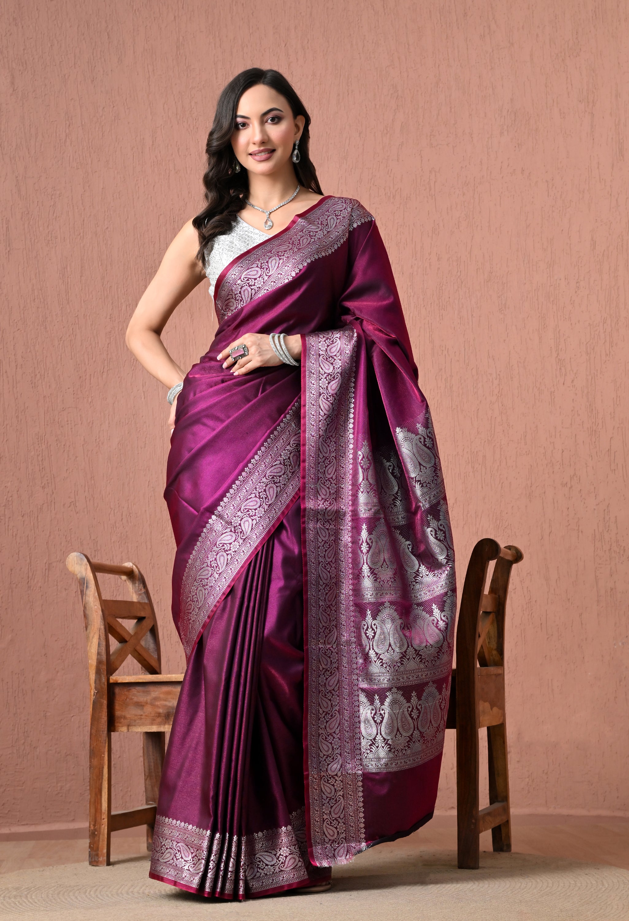 A woman modeling a luxurious wine-purple pure silk Banarasi saree featuring intricate silver zari paisley borders and a detailed pallu, paired with a silver-white sleeveless blouse.
