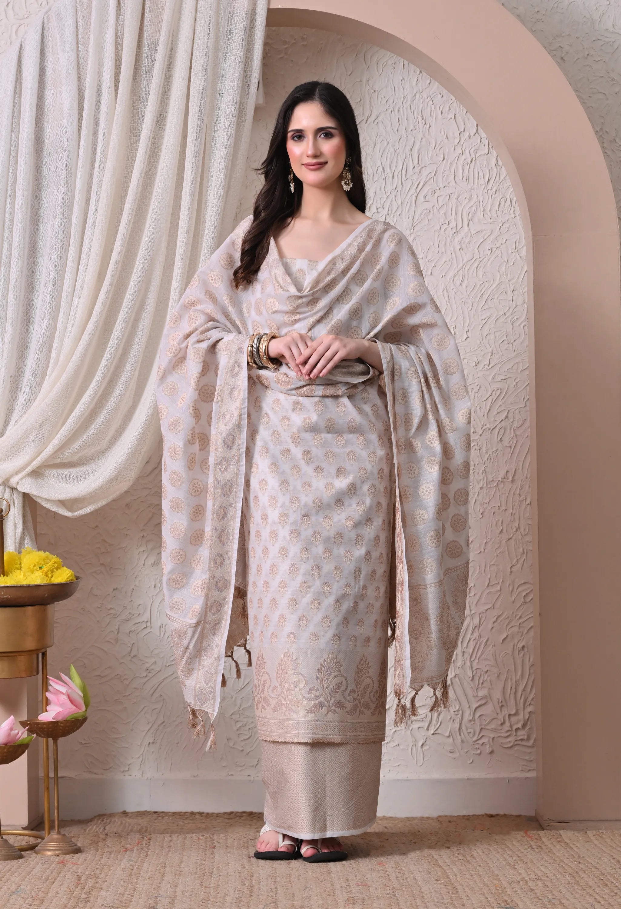 A woman posing in an elegant off-white and gold woven silk salwar suit set featuring intricate ethnic motifs, a detailed paisley border, and a matching tasseled dupatta.
