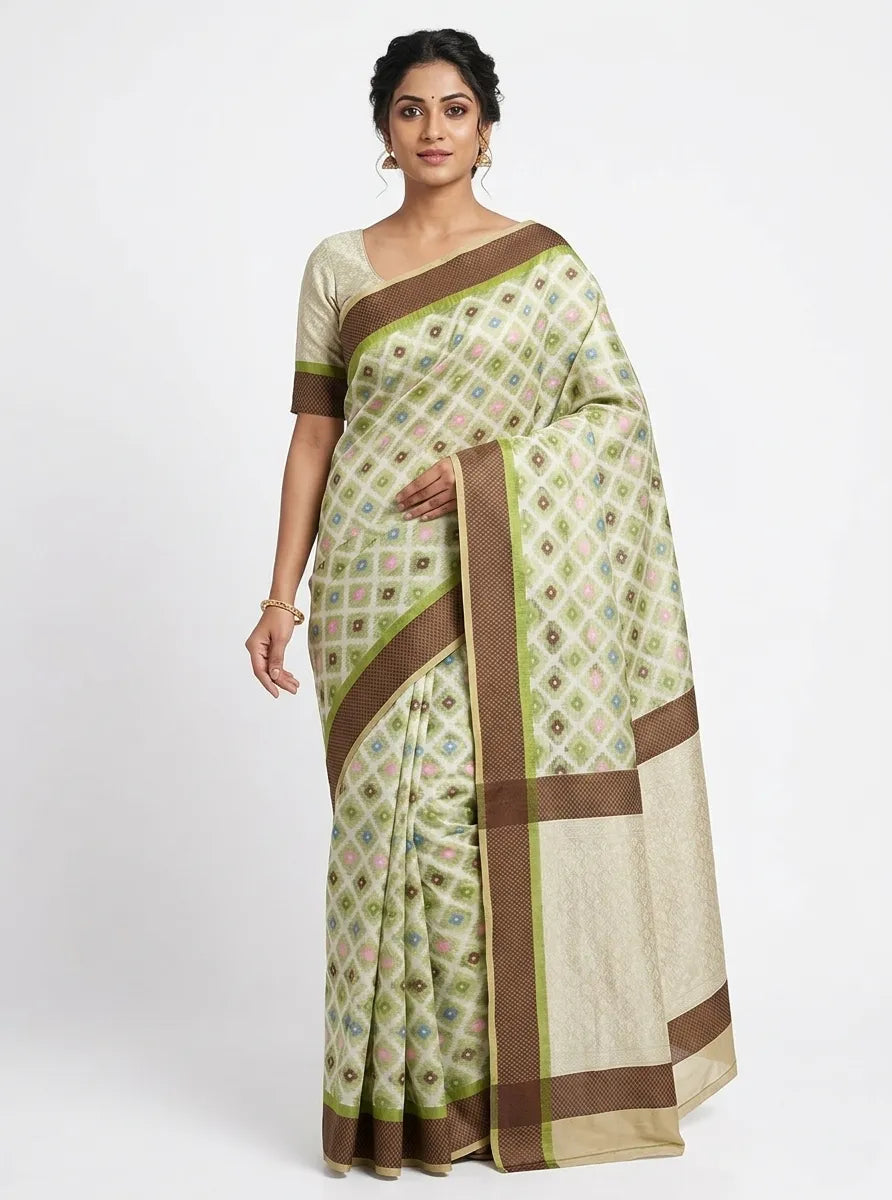 A woman posing in a sage green and cream Chanderi silk blend saree featuring a geometric diamond print. The saree has a wide, contrasting chocolate brown border with subtle gold zari work. She is wearing a matching sage green blouse and traditional gold jhumka earrings, set against a plain white backgroun