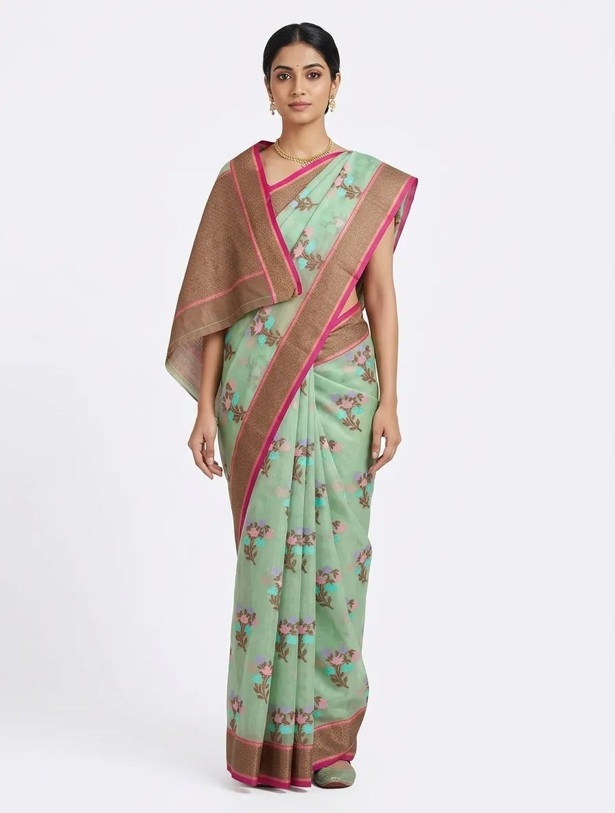 An Indian woman posing in a sea green floral printed saree with a wide bronze zari border and pink accents. She is wearing a gold necklace and earrings, standing against a plain white background.
