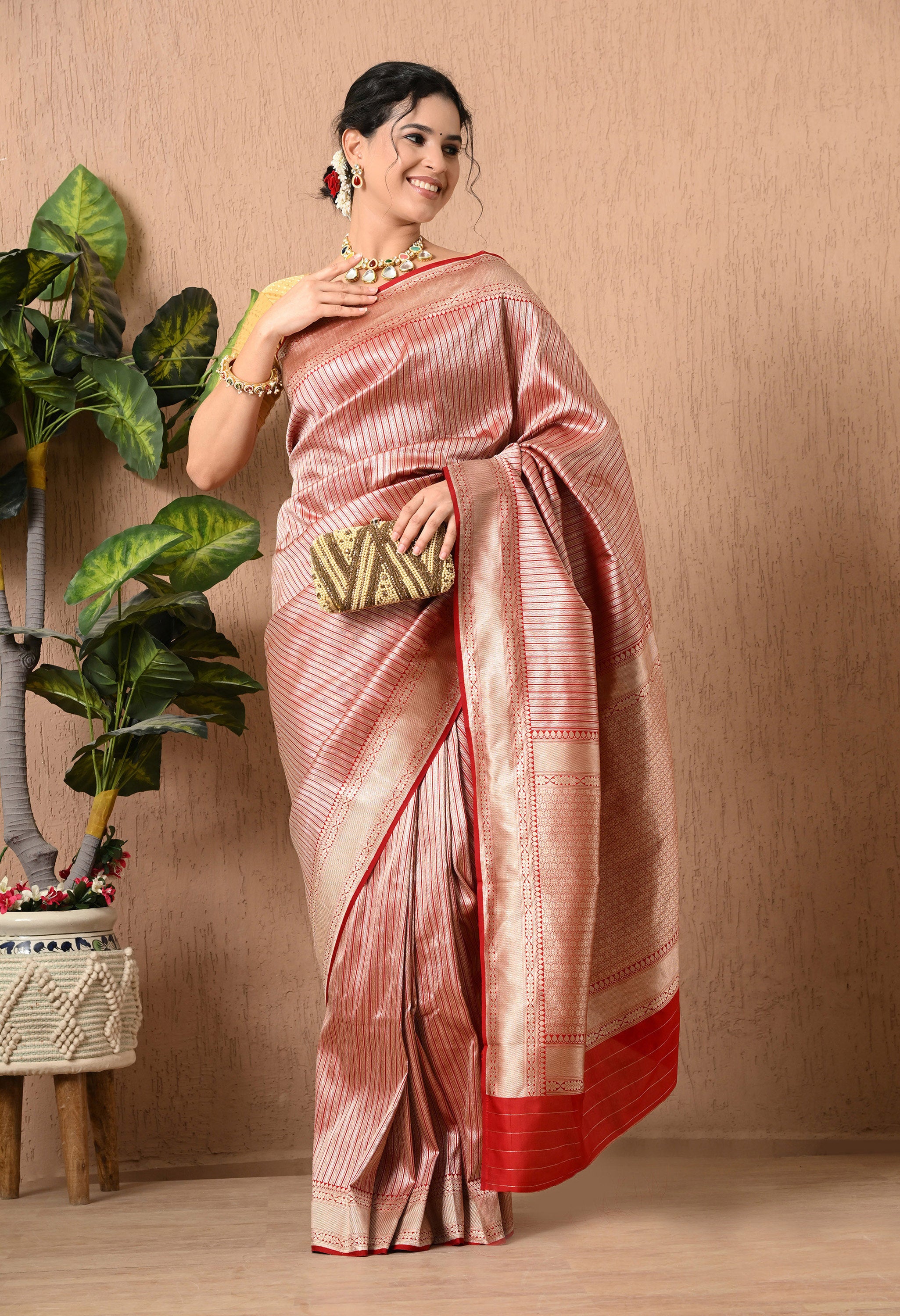 A full-length front view of a smiling woman posing in an elegant, Silk Mark Certified saree from The Weavers Shop by Raavi. The saree is crafted from premium Pure Katan Silk in a shimmering metallic red, gold  and silver tone, featuring sophisticated hand-woven vertical zari stripes throughout the body. She is holding a gold and brown patterned clutch and is styled with a mustard-gold textured blouse and a traditional kundan-style statement necklace.