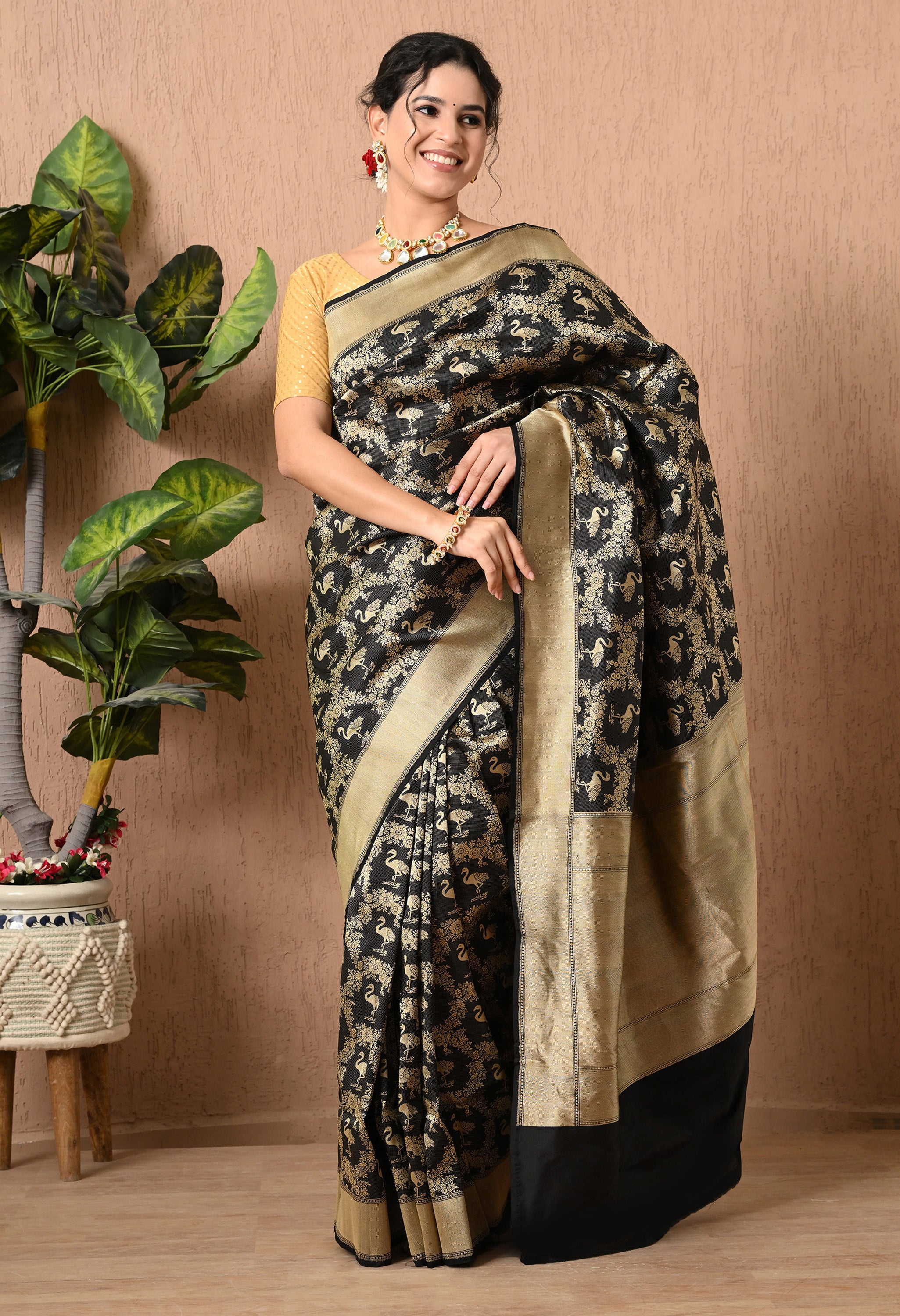 A full-length view of a woman smiling while modeling an elegant, black handwoven Katan Silk saree from The Weavers Shop by Raavi. The saree features a sophisticated Shikarga design, showcasing intricate handwoven gold zari work with detailed animal and floral motifs throughout the fabric. This Silk Mark Certified piece is finished with a wide, shimmering gold border and paired with a gold blouse and a traditionalnecklace.