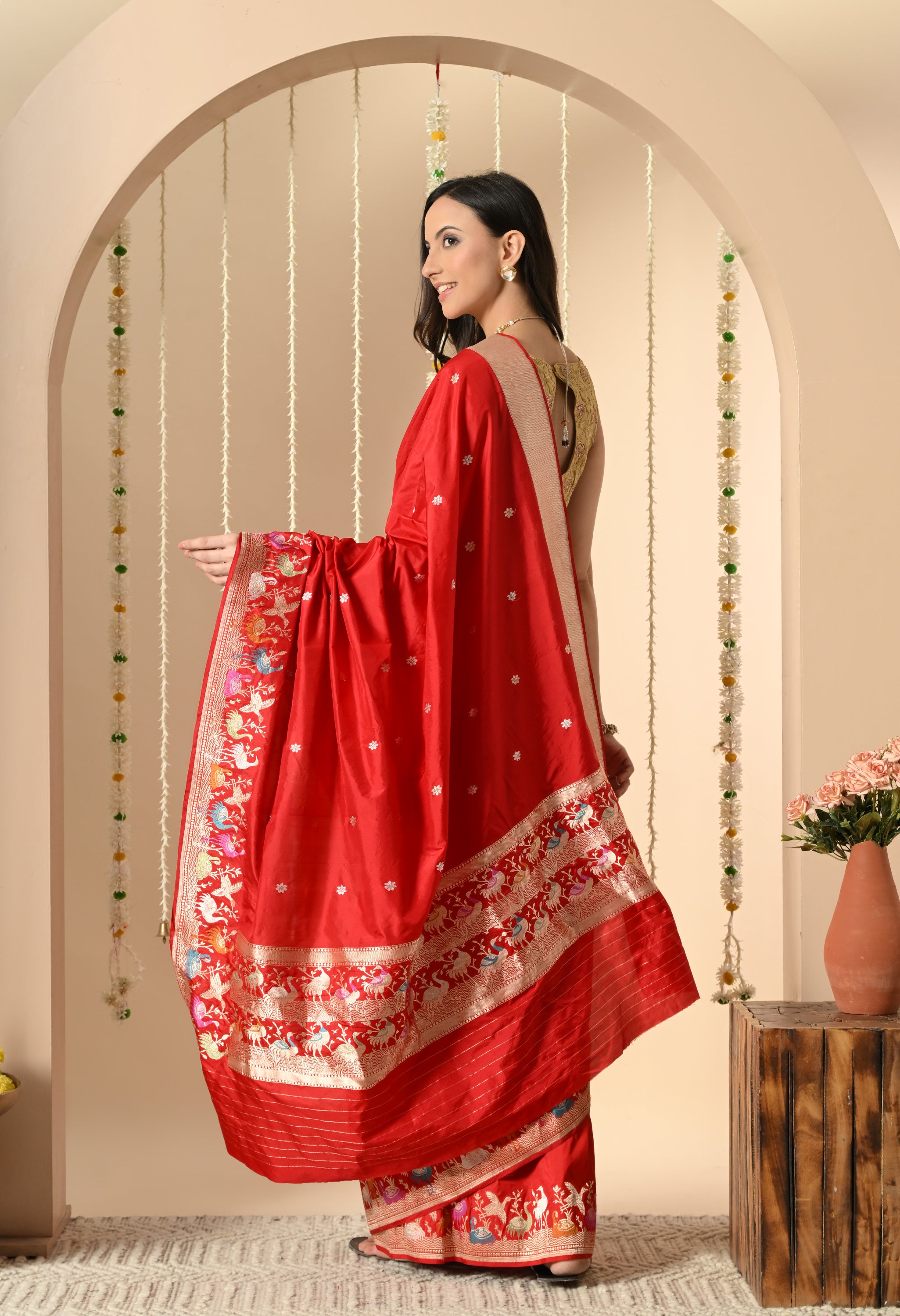 Modeling a Silk Mark certified red silk saree from The Weavers Shop by Raavi, a woman is shown in a three-quarter profile. The saree's focal point is the detailed, multicolored Shikarga border, featuring intricate woven motifs of animals and hunting scenes, complemented by a gold blouse.