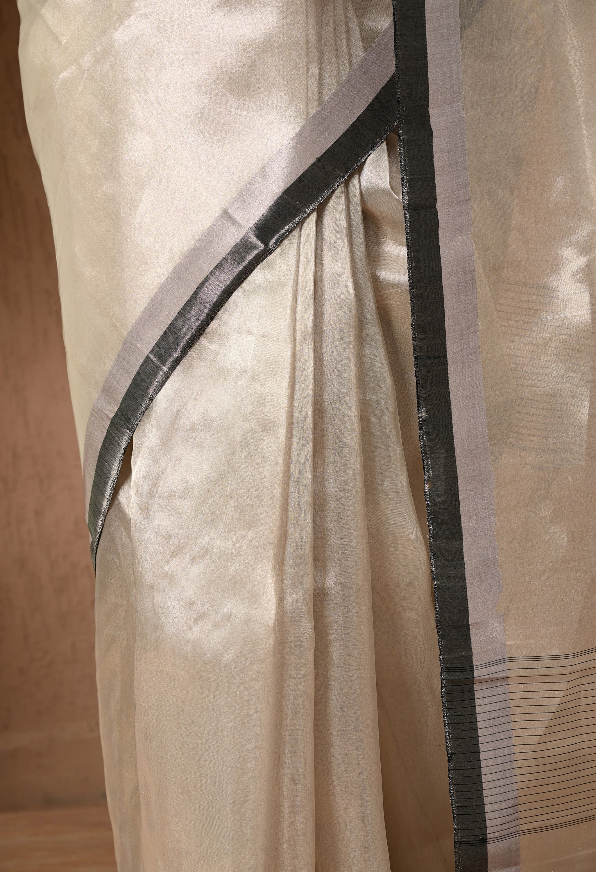 A close-up shot of the pleats and border of a silver tissue silk saree from The Weavers Shop by Raavi. The image highlights the fine, metallic texture of the semi-transparent tissue fabric and the contrasting black and silver border with subtle horizontal striped detailing. The luminous fabric is neatly draped, showcasing its high-shine finish and delicate weave.