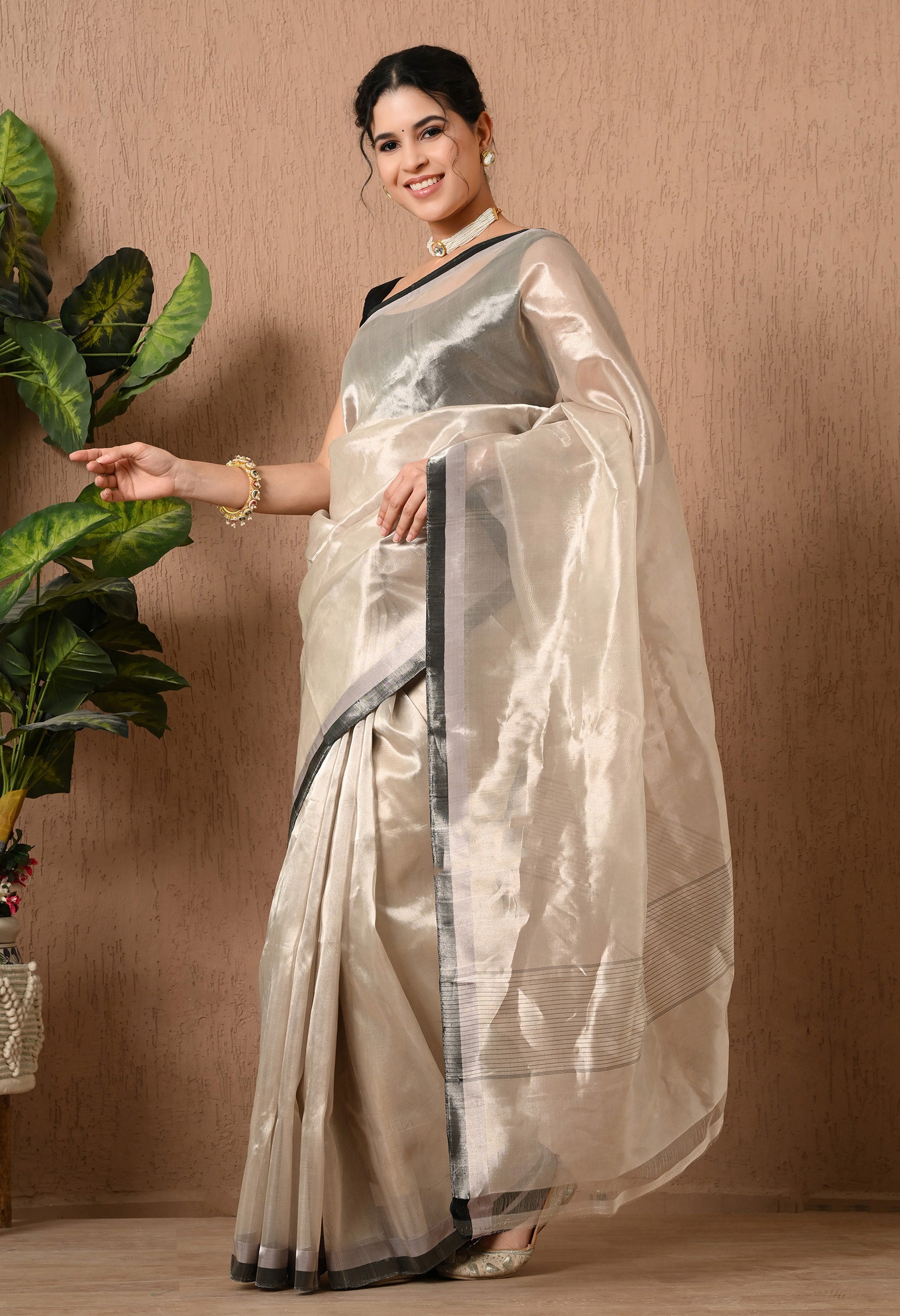 A side-profile view of a woman wearing a shimmering silver tissue silk saree by The Weavers Shop by Raavi. The saree features a luminous, semi-transparent fabric with a high-shine metallic finish and a contrasting black and silver border. She is styled with a sleeveless black blouse, a pearl choker, and traditional bangles, standing gracefully against a textured beige wall with a green plant to her side.