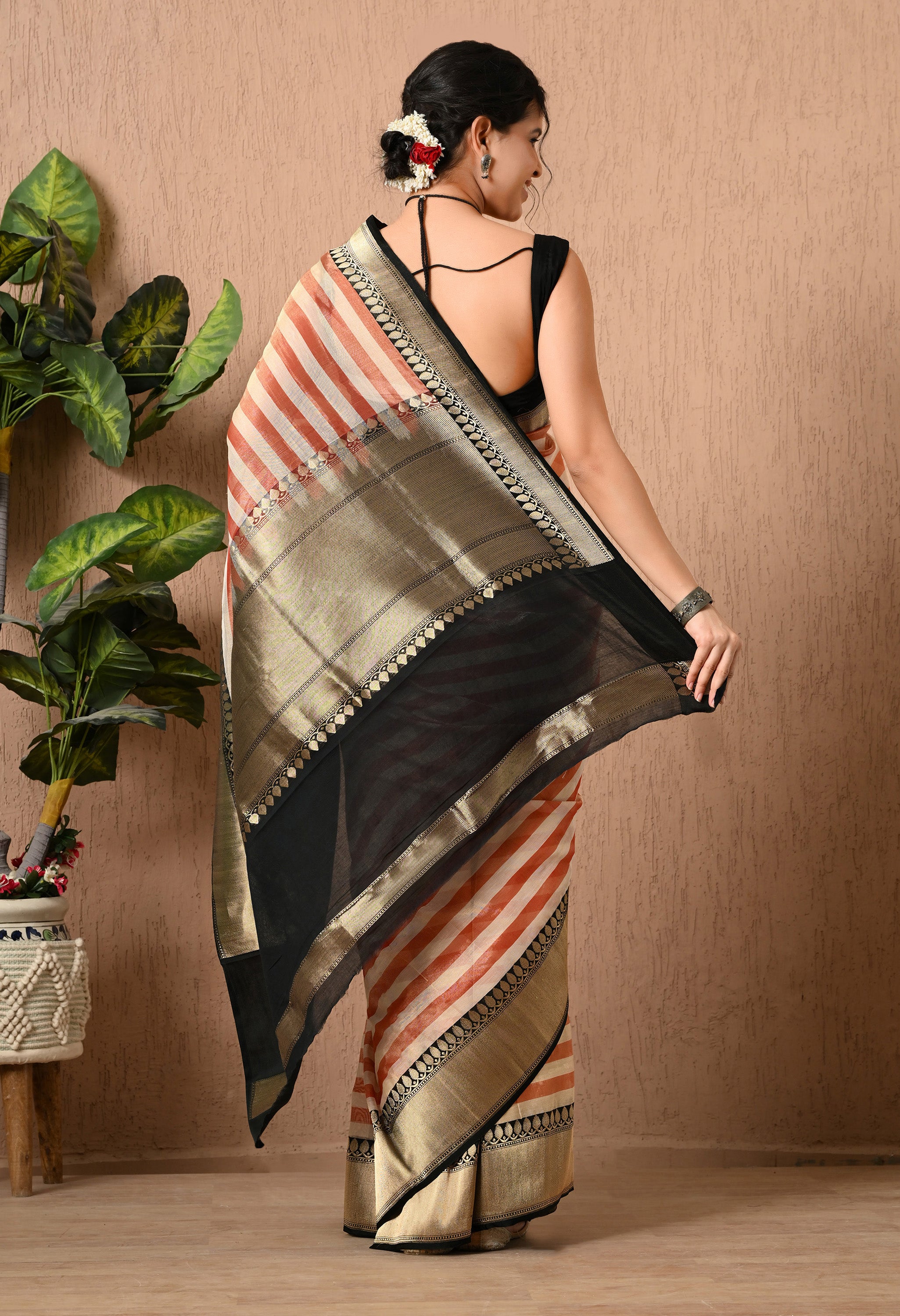 Back view of a woman showcasing a handwoven Tissue Silk saree by The Weavers Shop by Raavi. The image highlights the striped pattern in rust-red and cream, featuring a prominent black pallu with thick gold zari borders and intricate traditional motifs. She is wearing a black  blouse with and has her hair styled in a bun 