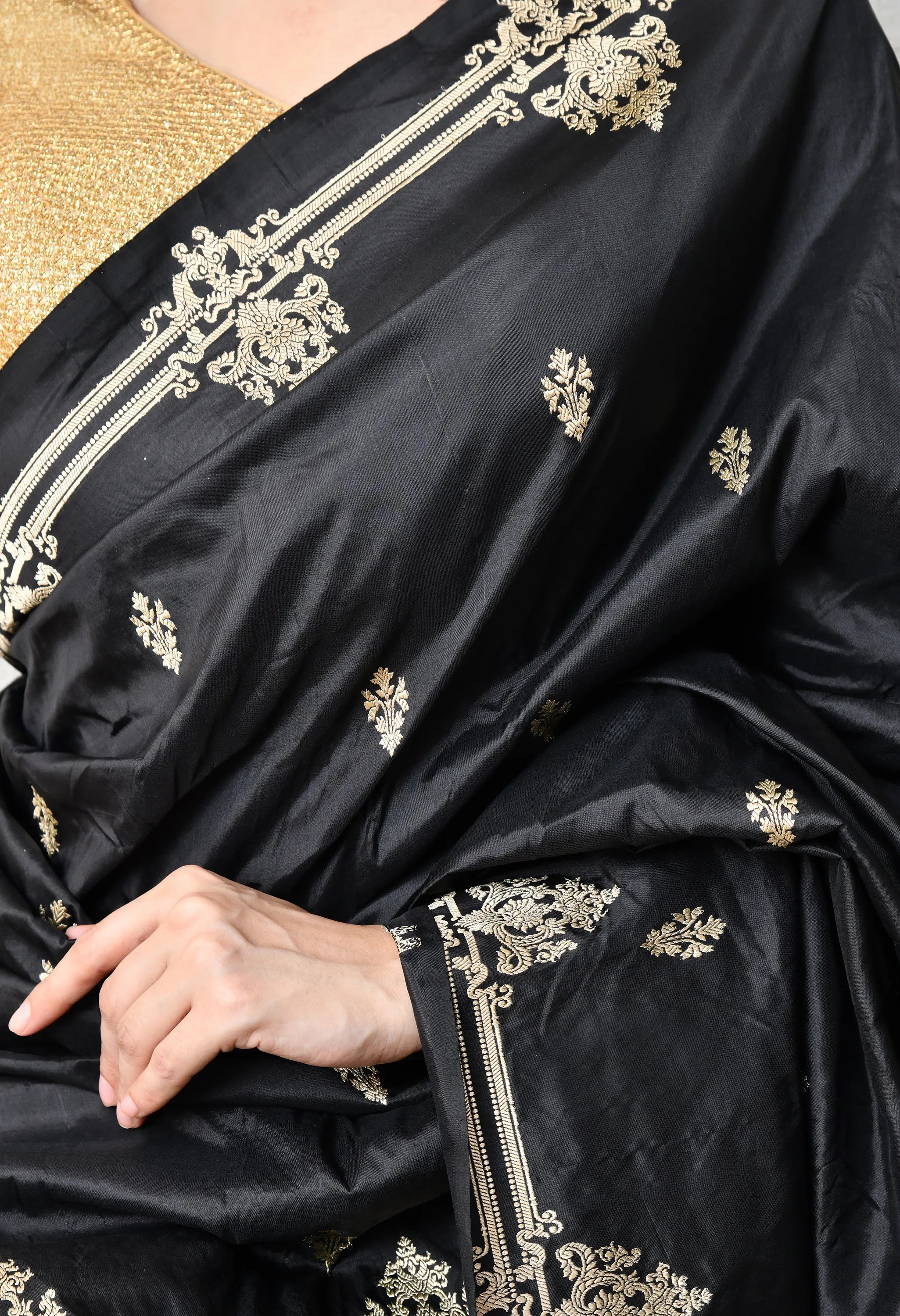 A close-up of a woman wearing a Silk Mark Certified black Katan silk saree from The Weavers Shop by Raavi. The saree has intricate gold zari work, featuring large, stylised gold motifs on the shoulder pleats and a thin gold border.
