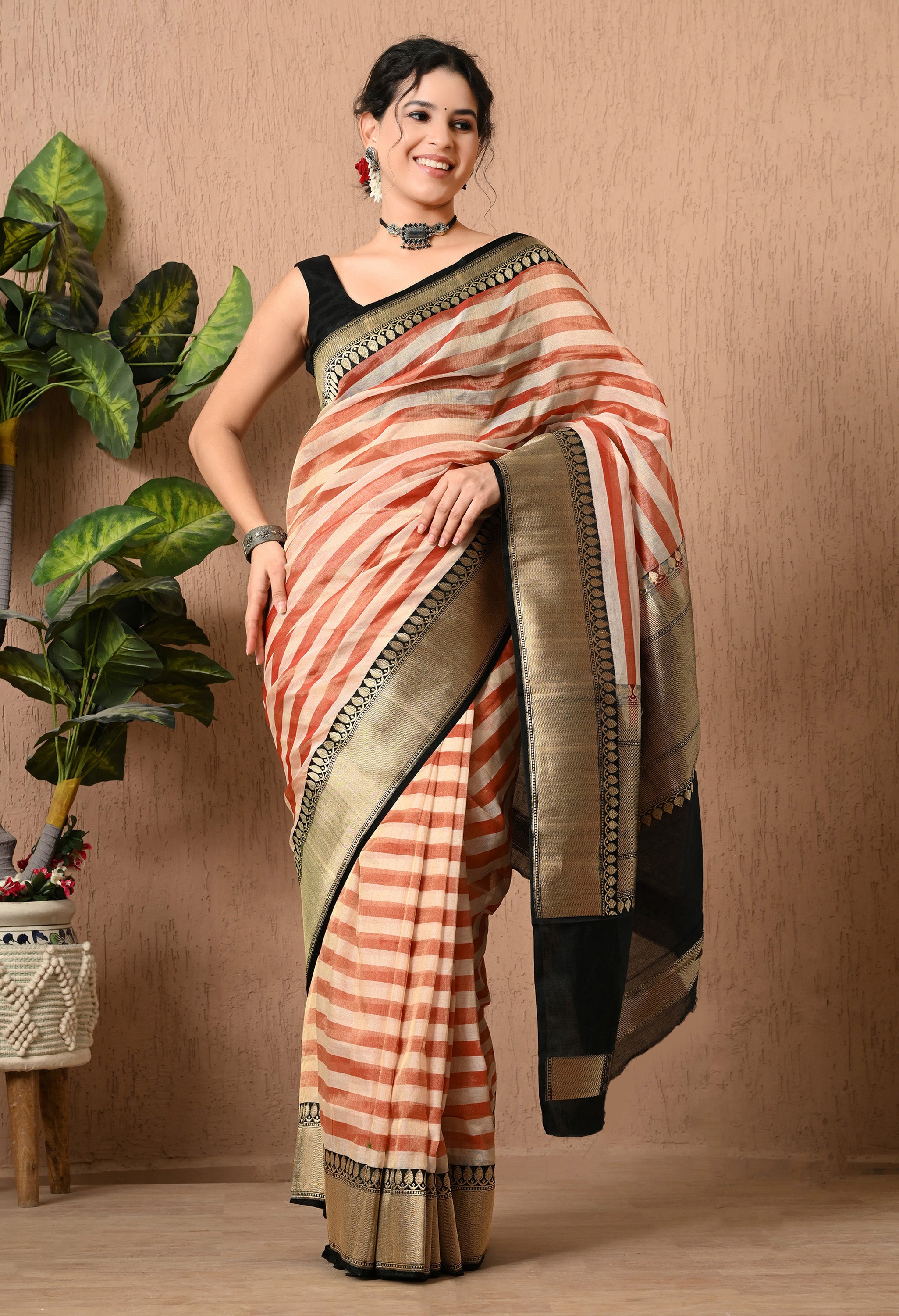 A woman posing in a The Weavers Shop by Raavi saree made of handwoven Tissue Silk. The saree features a horizontal striped pattern in rust-red and cream, complemented by a wide black border and pallu decorated with intricate gold zari work. She is wearing a sleeveless black blouse and oxidized silver jewelry, standing against a neutral textured wall.