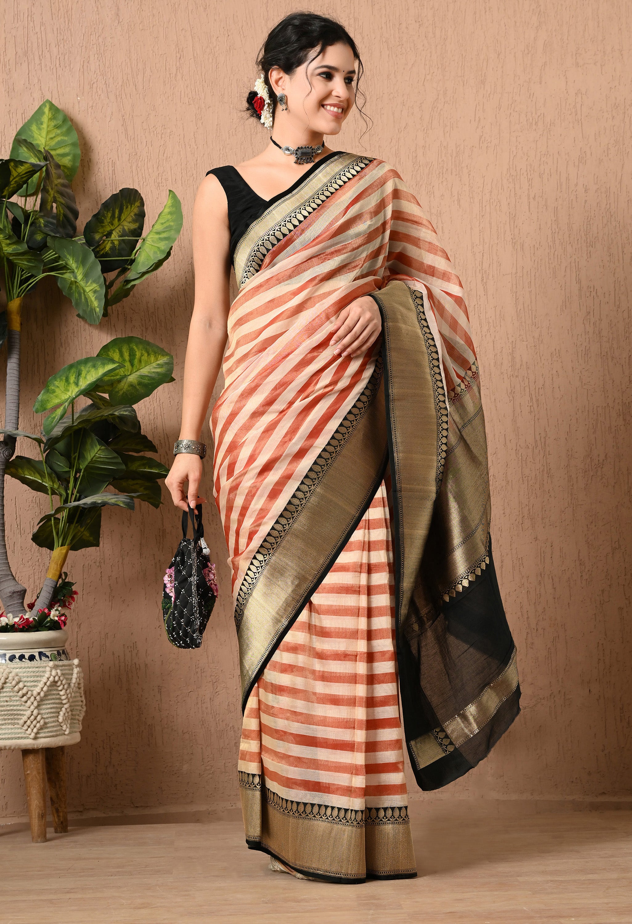 A woman smiling and posing in a handwoven Tissue Silk saree from The Weavers Shop by Raavi. The design features a rust-red and cream striped pattern paired with a thick black border and pallu showcasing intricate gold zari work. She is holding a small potli bag in her right hand and is styled with a black blouse and jewelry. She stands against a textured beige wall with a potted green plant nearby.