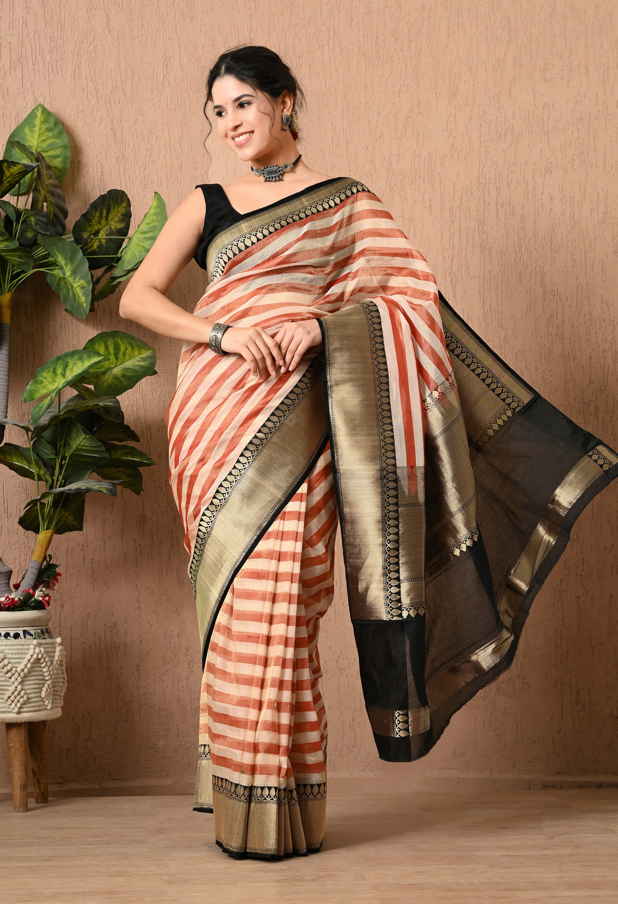 A dynamic shot of a woman smiling and posing in a handwoven Tissue Silk saree from The Weavers Shop by Raavi. The black pallu is captured mid-air, flying out to the side to showcase the weightless quality of the fabric and the detailed gold zari work. The saree features a rust-red and cream striped pattern, paired with a black sleeveless blouse and oxidized silver jewelry.