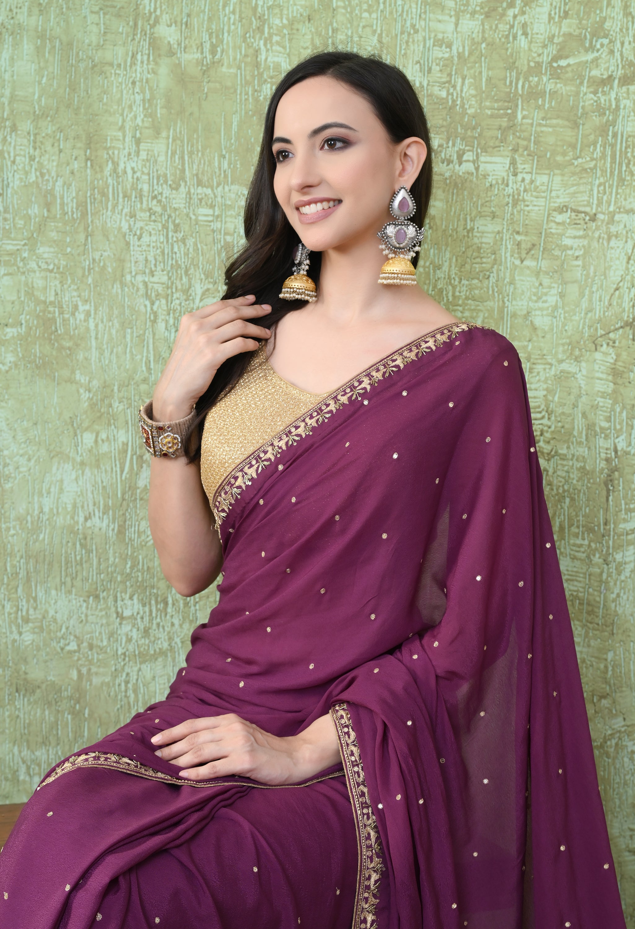 A close-up portrait of a woman wearing a wine-colored crepe saree from The Weavers Shop by Raavi. The shot captures the fine detail of the gold zardozi hand-embroidery along the neckline and the subtle sparkle of the sequin work on the fabric. She wears ornate traditional earrings and a gold sleeveless blouse.