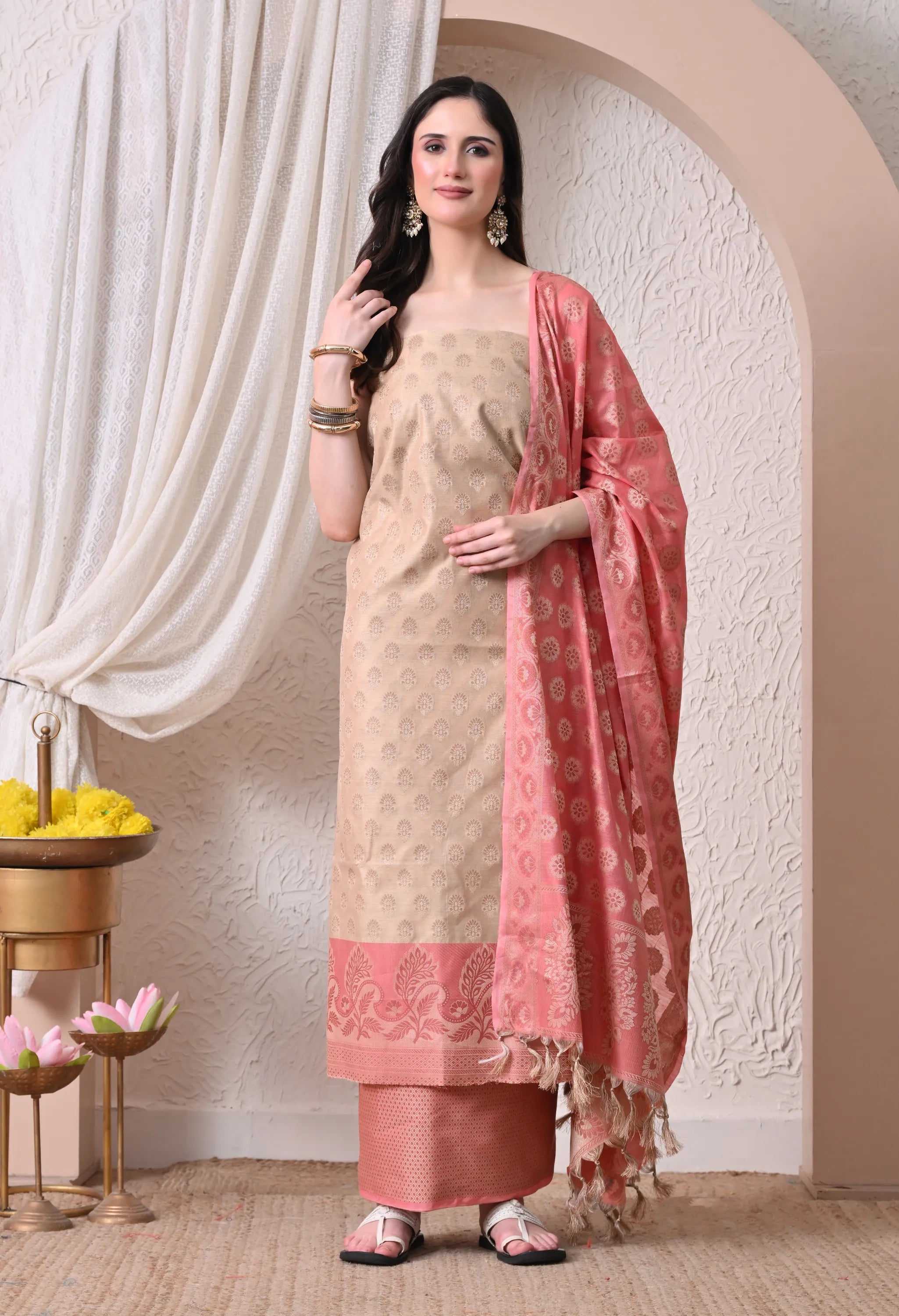A woman posing in an elegant beige and pink woven silk salwar suit set featuring detailed ethnic motifs, a pink contrast paisley border, and a matching tasseled dupatta.
