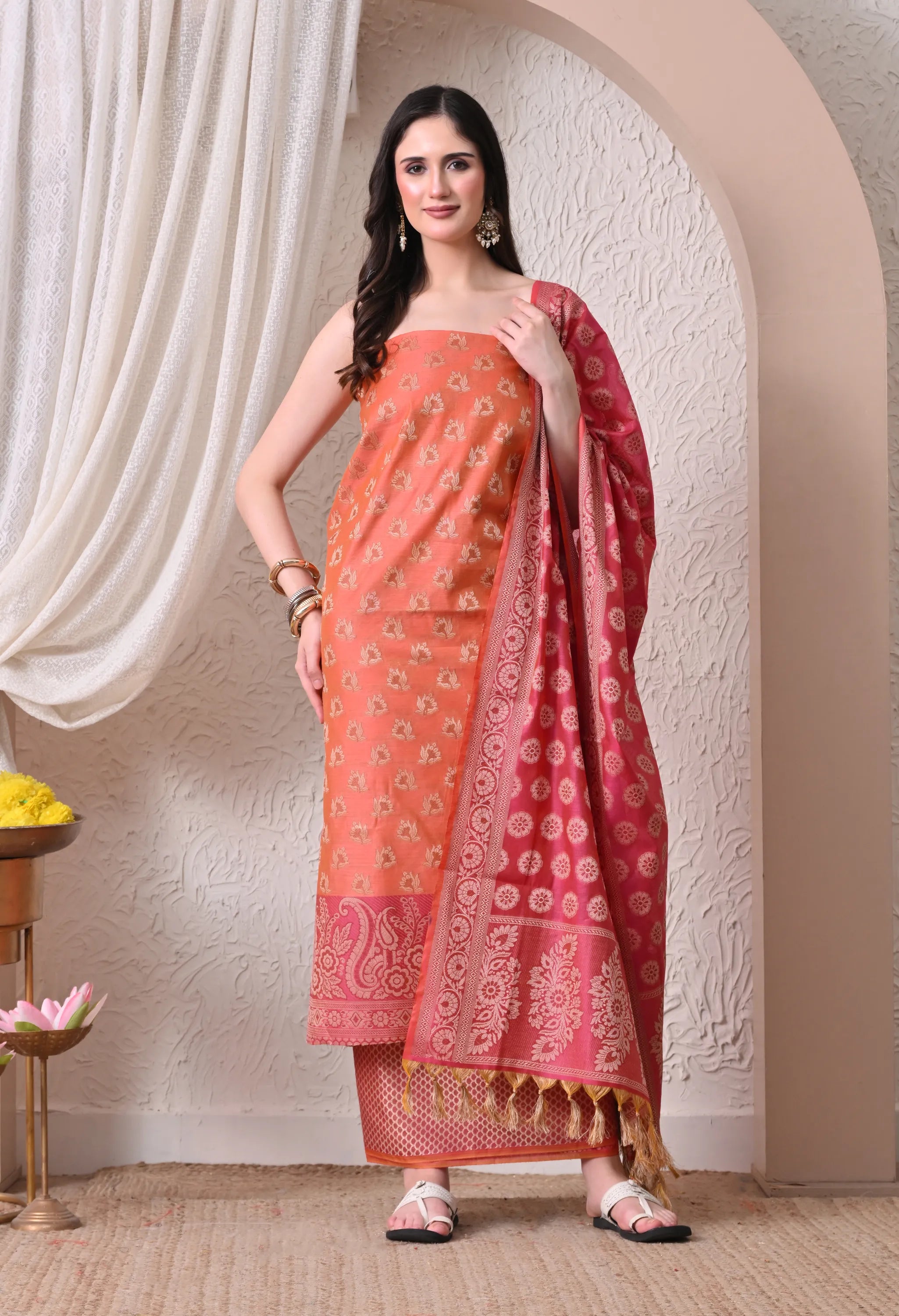 A woman posing in a vibrant peach and magenta woven silk salwar suit set featuring detailed ethnic motifs, a magenta paisley border, and a matching tasseled dupatta.

