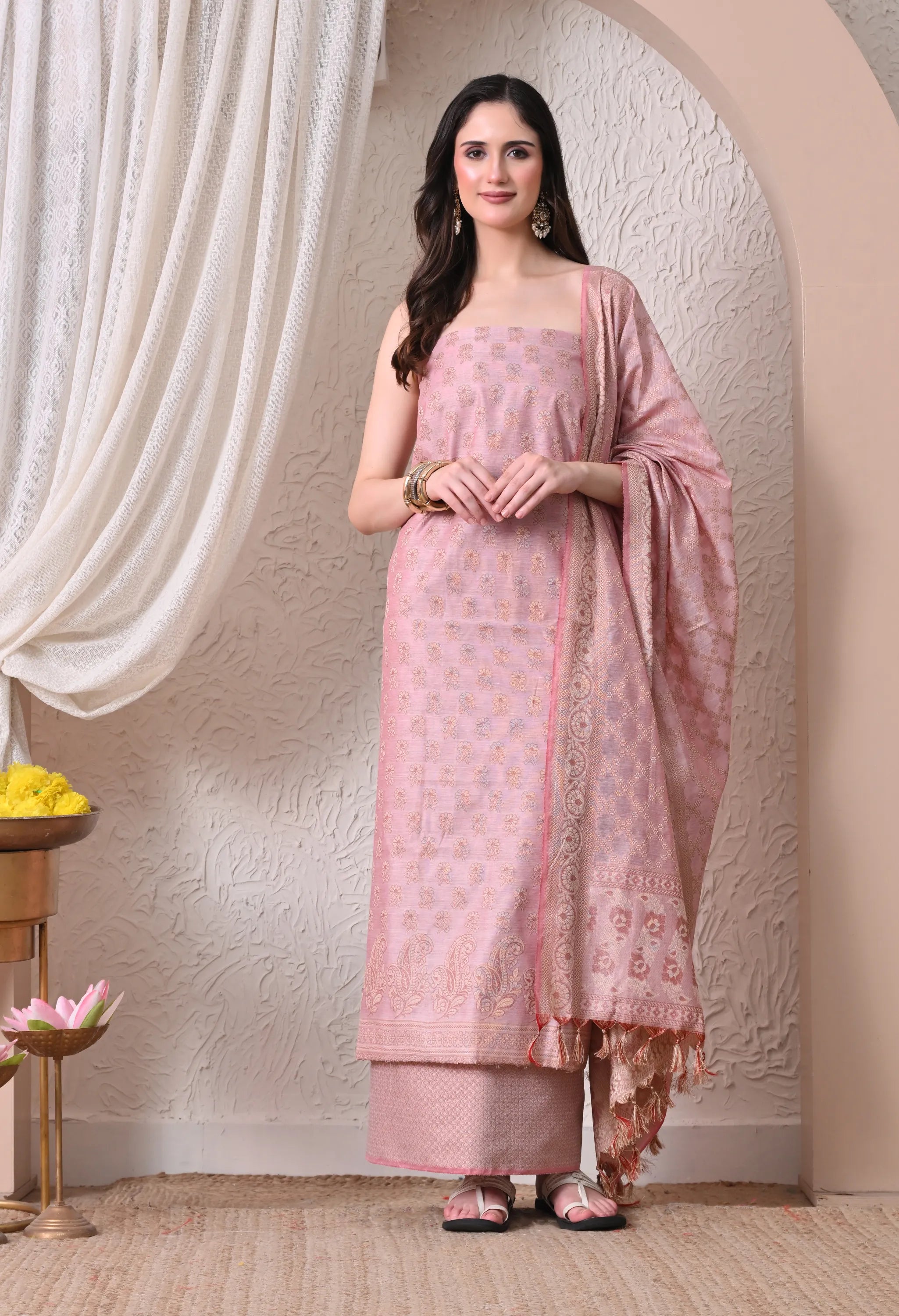 A woman posing in an elegant onion pink and gold woven silk salwar suit set featuring intricate ethnic motifs, a detailed paisley border, and a matching tasseled dupatta.
