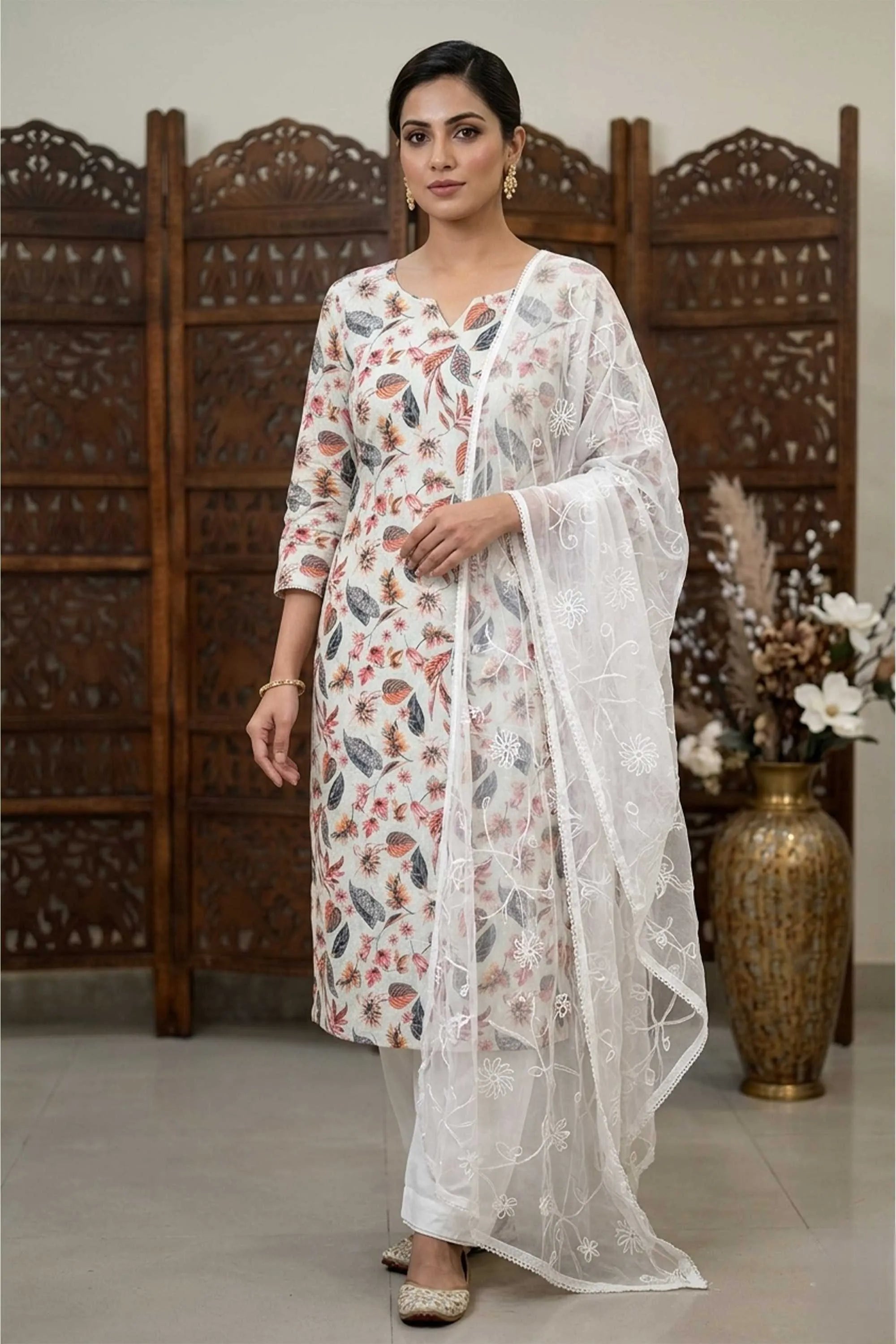 Woman wearing a white floral print kurta set with a matching sheer white dupatta, standing against a carved wooden screen backdrop.