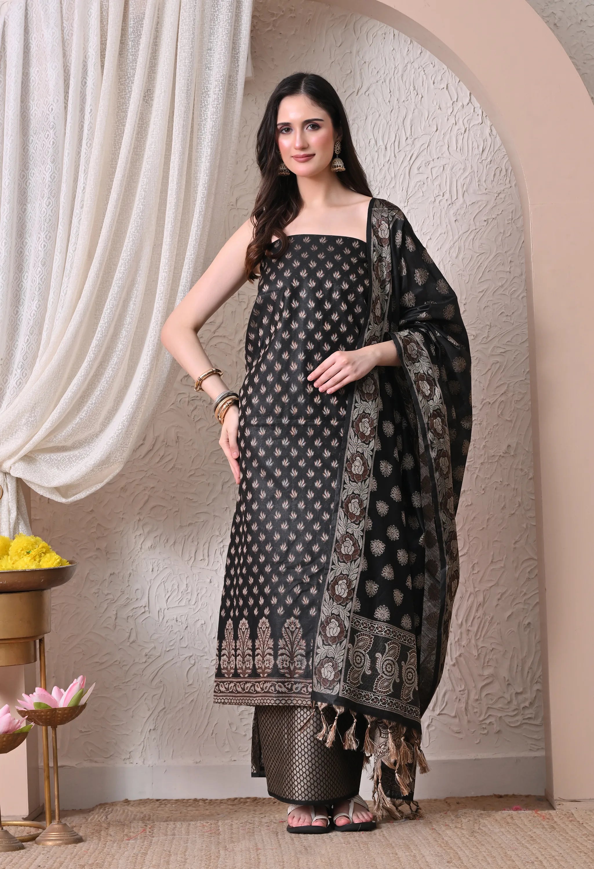 A woman posing in a luxurious black and gold woven silk salwar suit set featuring detailed leaf motifs, an ornate traditional border, and a matching tasseled dupatta.
