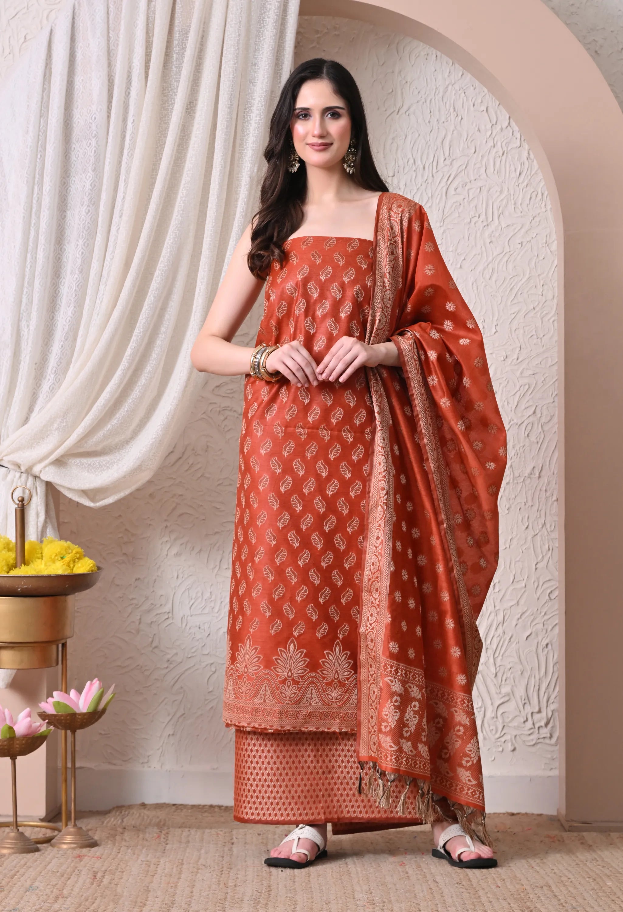  A woman posing in a vibrant rust orange and gold woven silk salwar suit set featuring detailed leaf motifs, an ornate traditional border, and a matching tasseled dupatta.
