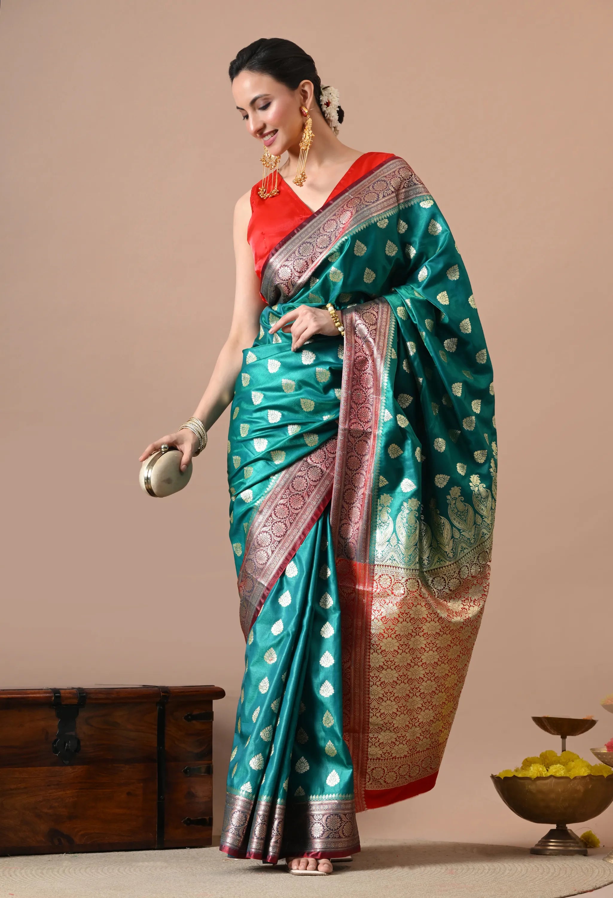 Woman modeling a teal silk blend saree with gold floral motifs, a red and gold woven border, and a matching red blouse.
