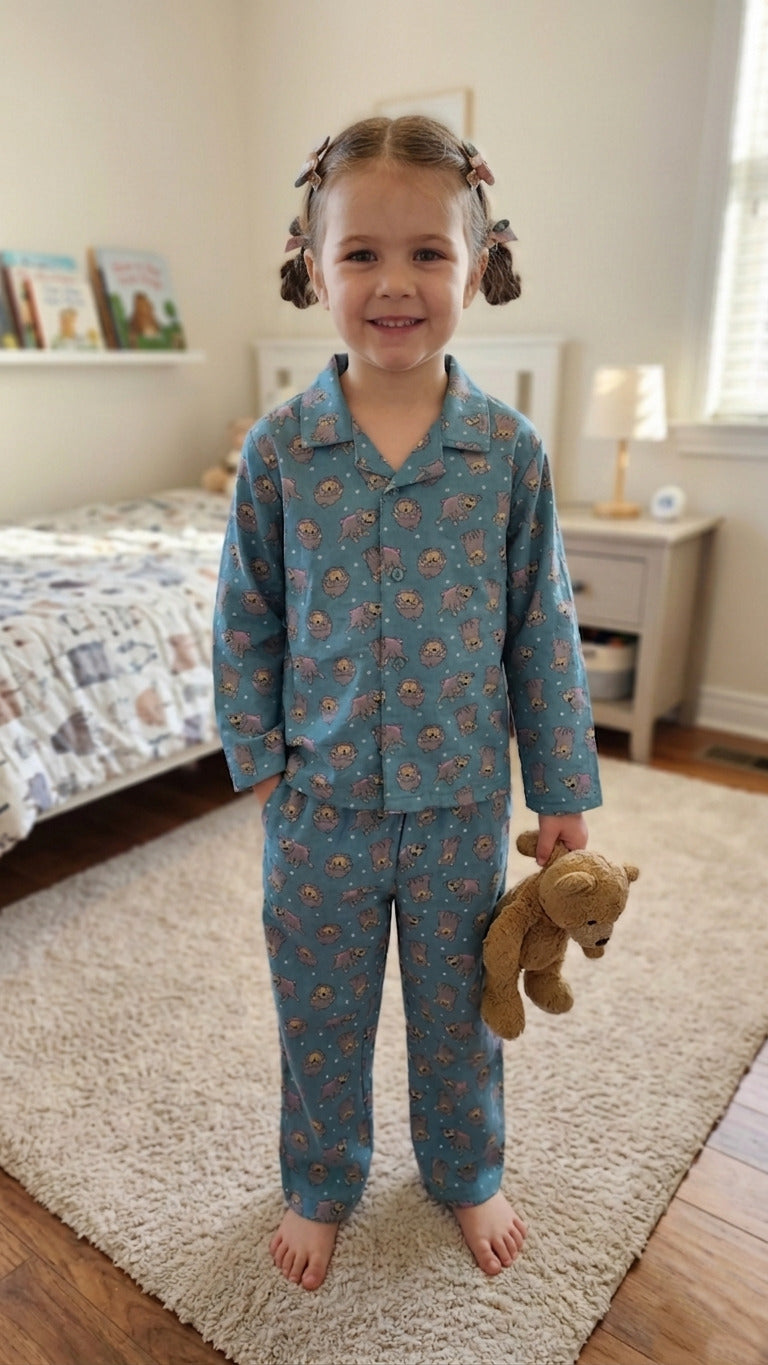 A young girl smiling in a bedroom, wearing a teal blue cotton night suit with a grey hedgehog print and holding a teddy bear.
