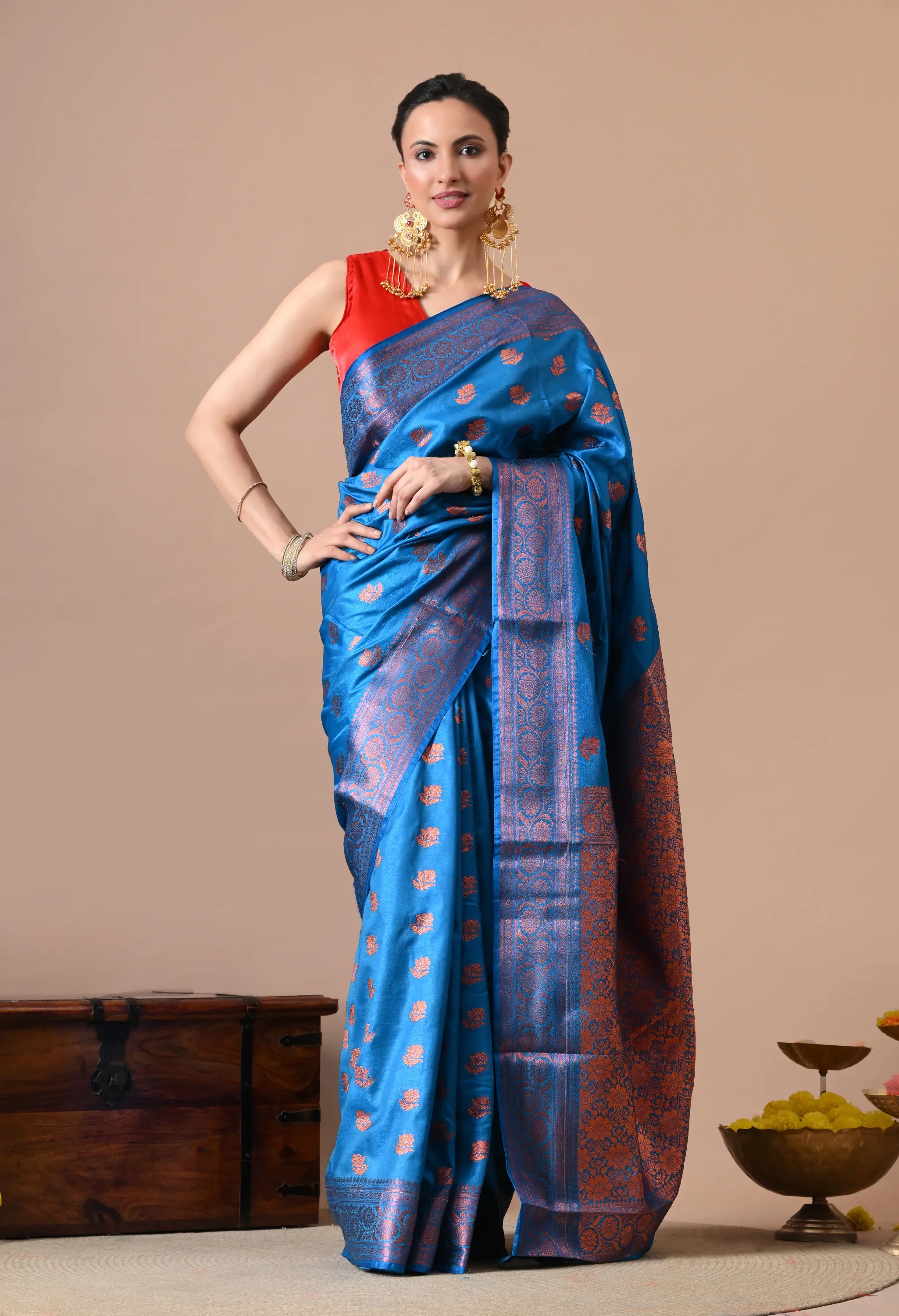 Woman in a peacock blue silk-blend saree with copper zari woven motifs and border, paired with a red sleeveless blouse and heavy gold earrings.
