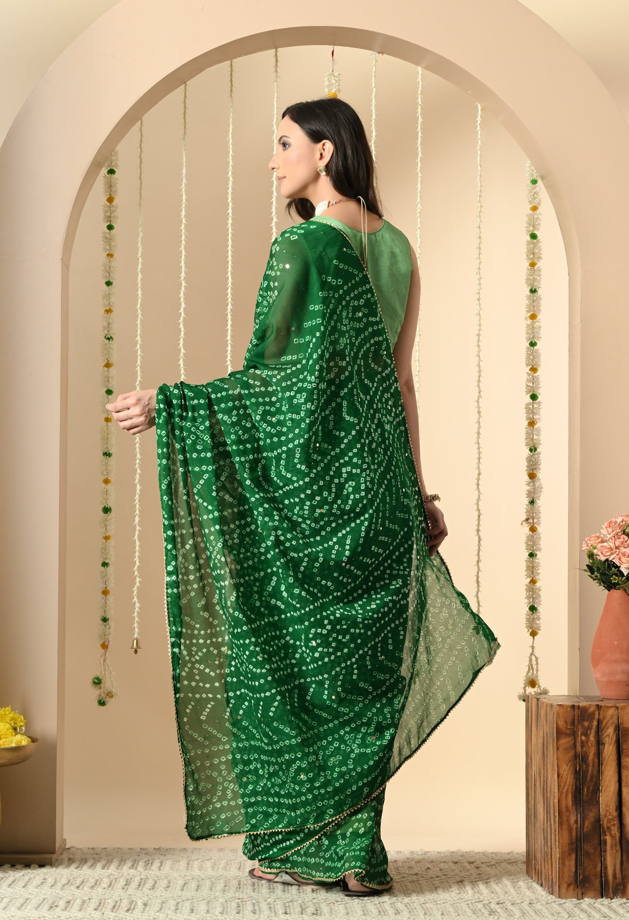 A back view of a woman showcasing the lightweight and fluid drape of the green Handcrafted Bandhani Georgette Saree from Rajasthan. The image emphasizes the overall repeating tie-dye motif and the saree's elegant fall, paired with a green sleeveless blouse.