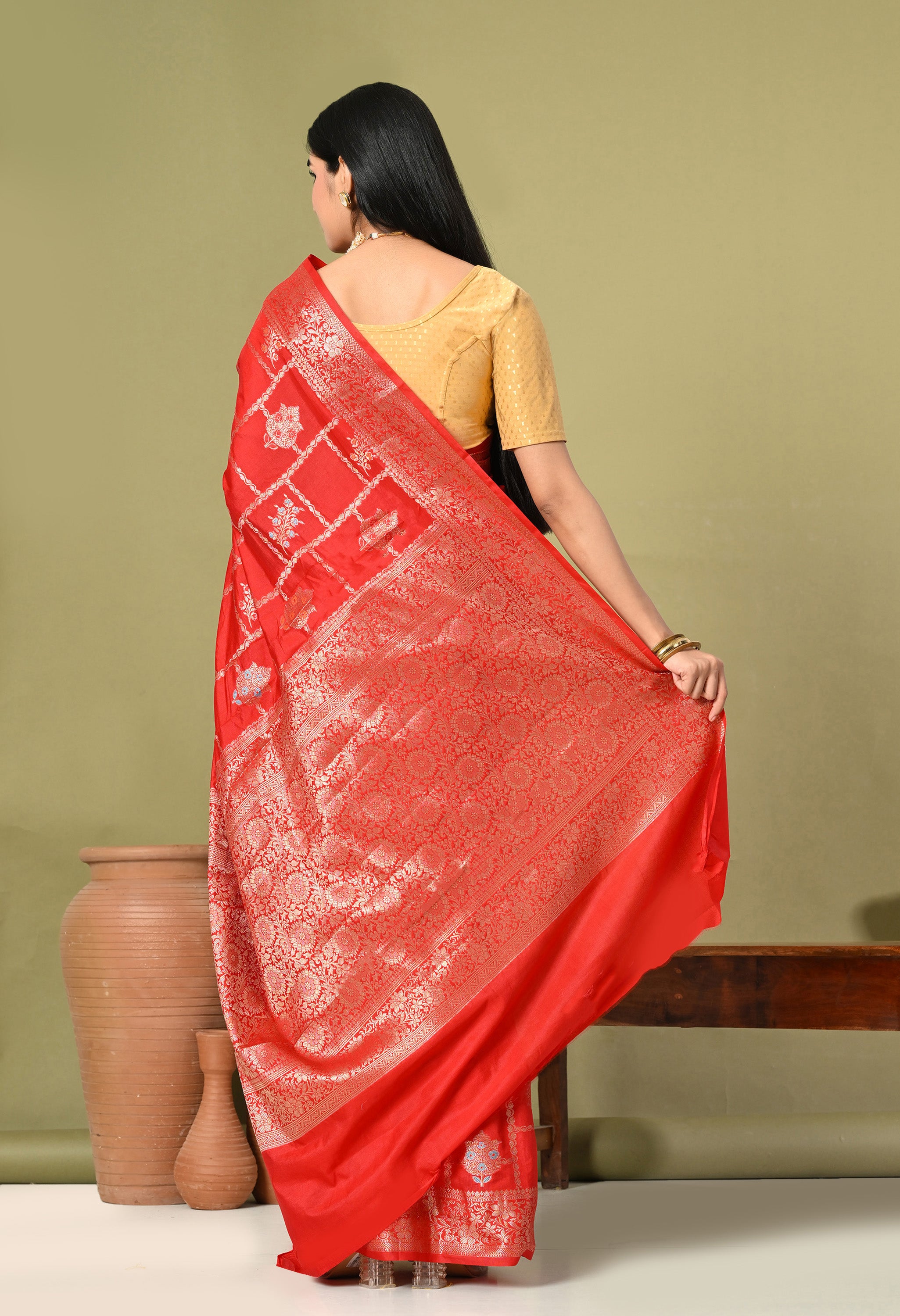 A back view of a woman showcasing a vibrant red Banarasi silk saree featuring exquisite Tilfi work. She holds the pallu (decorative end) outward to reveal a dense, intricate gold zari pattern consisting of traditional floral and vine motifs.

The main body of the saree displays a grid-like pattern with alternating silver and gold floral butis. She wears a contrasting gold textured blouse with a deep round back. The background is a muted olive green, complemented by rustic terracotta vases and a wooden benc