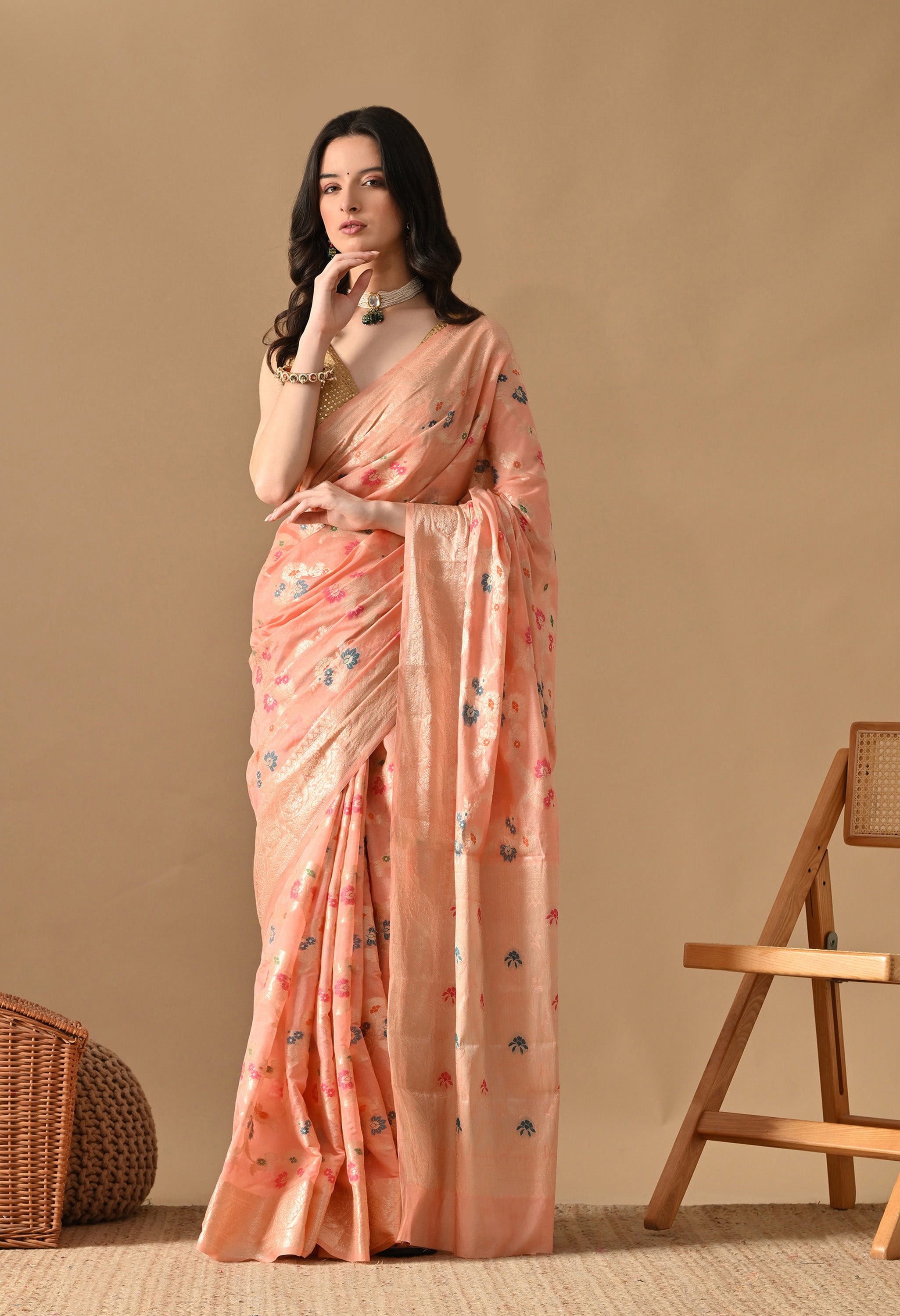 A woman stands against a beige background, elegantly draped in a peach-coloured silk saree from The Weavers Shop by Raavi. The saree features intricate Tilfi work with Zari, showcasing delicate floral motifs in shades of blue and pink across the body. It is finished with a broad, shimmering gold zari border and pallu. She pairs the saree with a sleeveless gold sequined blouse, a pearl choker with a green emerald drop, and matching jewellery.