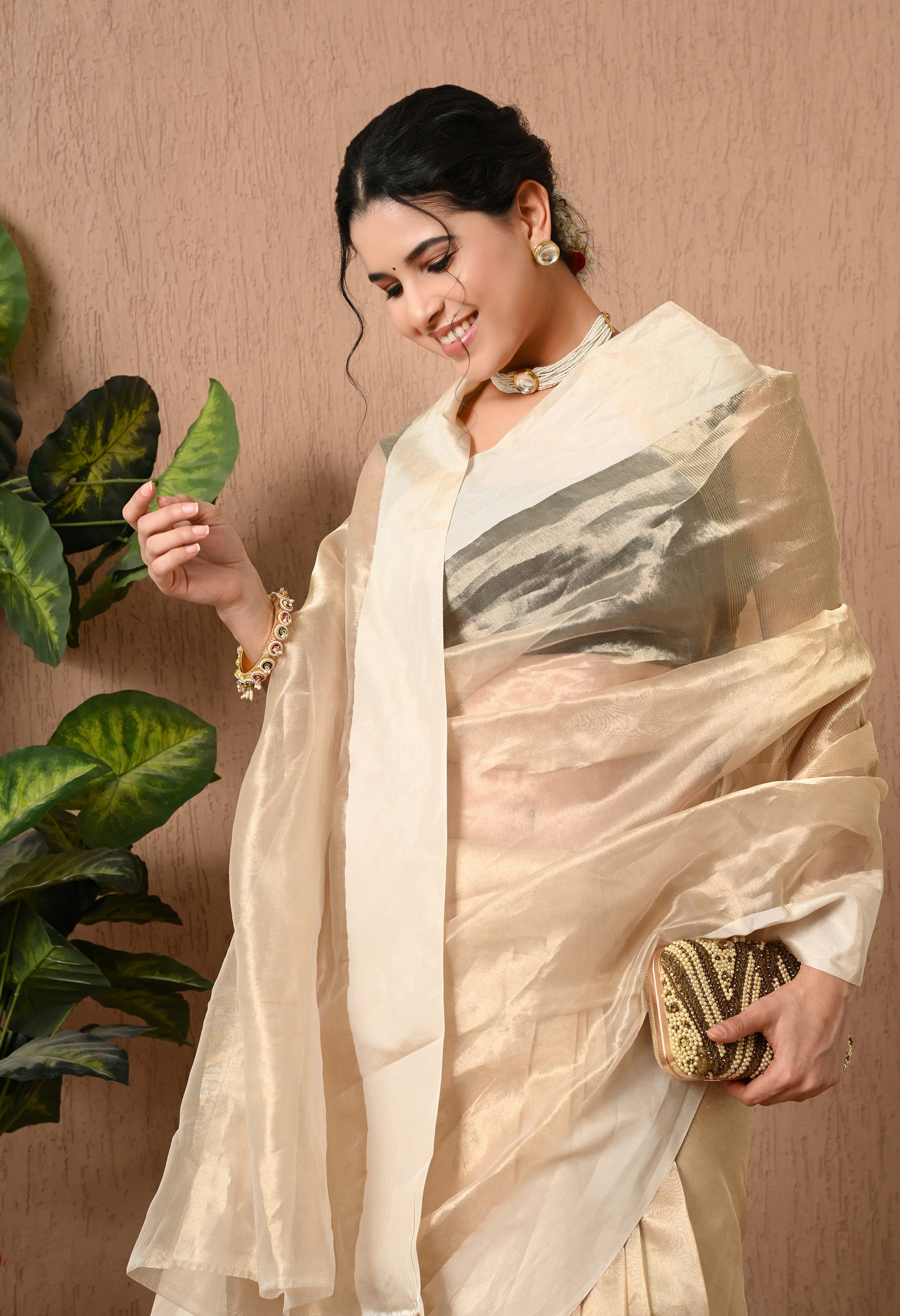 A close-up of a woman smiling and looking down, showcasing a shimmering champagne-gold tissue silk saree from The Weavers Shop by Raavi. The translucent pallu is draped over her shoulder and arm, layered over black blouse. She is wearing a choker, matching stud earrings, and a bangle, while holding a beaded gold and brown clutch.