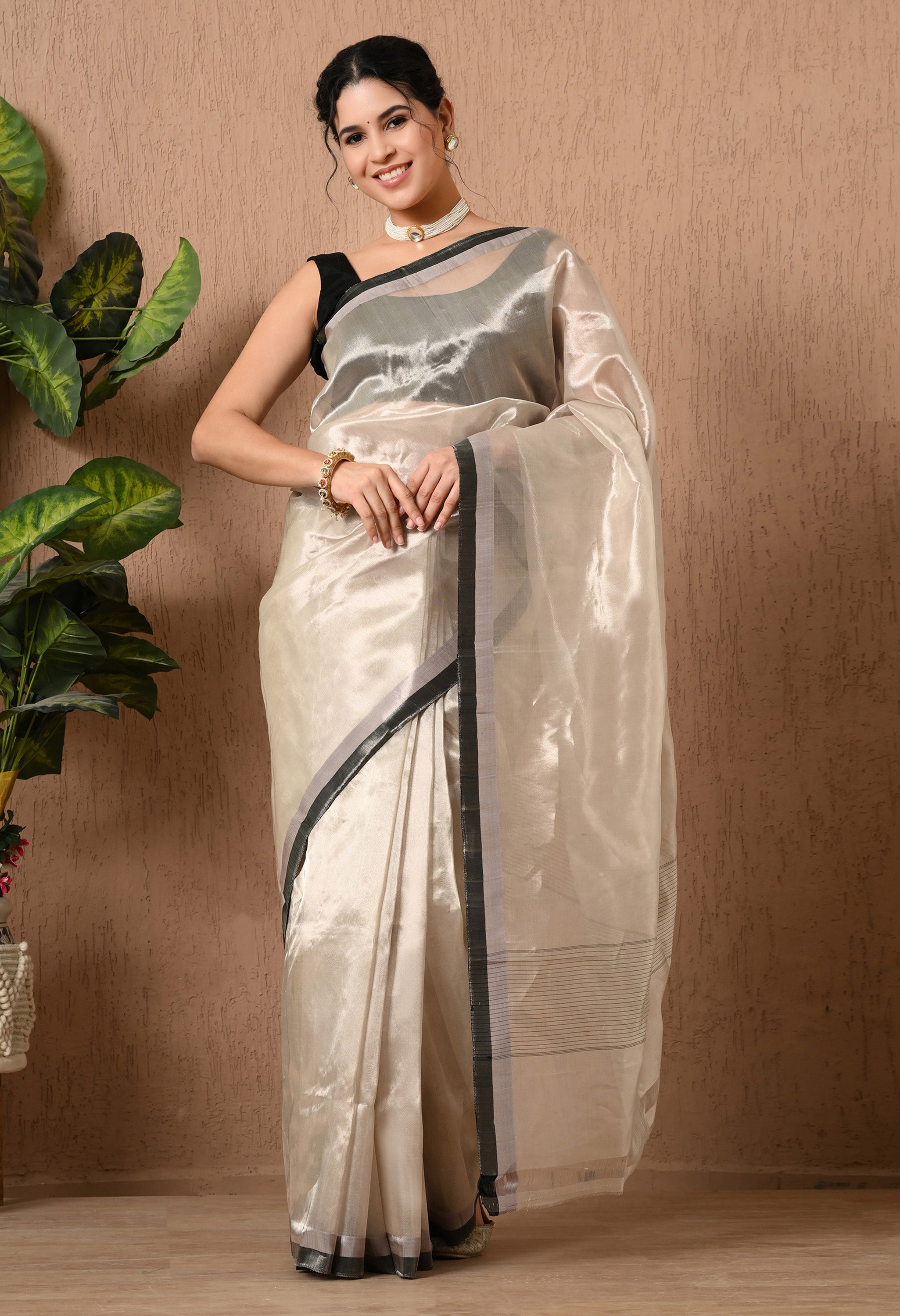 A woman poses in a shimmering silver tissue silk saree from The Weavers Shop by Raavi. The saree features a translucent, metallic sheen and a sleek black border with silver accents. She has paired it with a sleeveless black blouse, a white beaded choker, and matching earrings. The background is a textured beige wall with a large green leafy plant to the left.