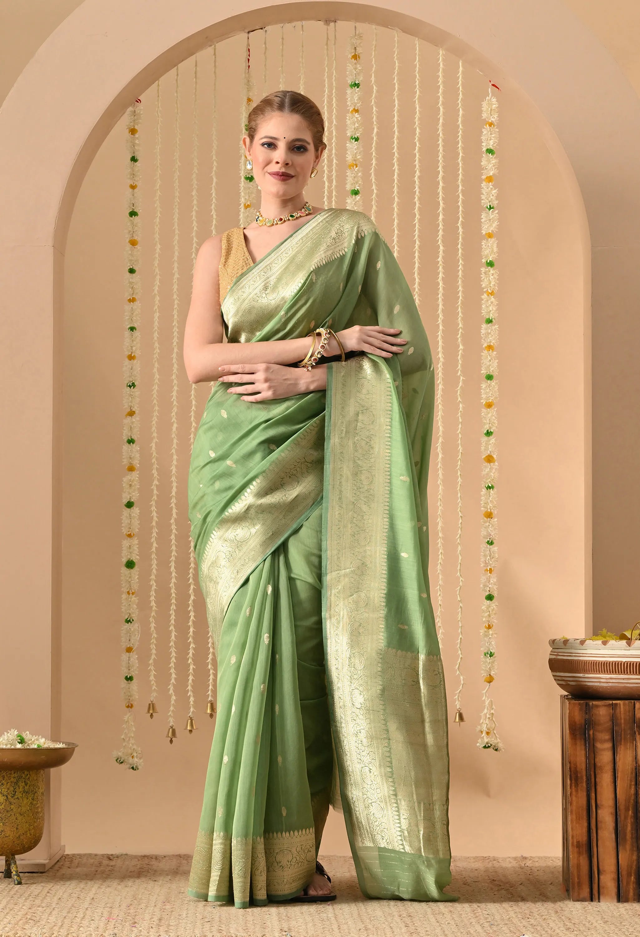 A woman stands elegantly under a floral archway, modeling a Silk Mark Certified Light Green Pure Silk Saree from The Weavers Shop by Raavi. This hand-woven Banarasi masterpiece features the prestigious Karuwa Weave technique, showcasing small, delicate zari work butis (motifs) scattered across the body. The saree is designed with a broad, shimmering gold zari border and a heavily detailed pallu, paired with a sleeveless gold  blouse. The soft, pastel green hue of the silk is highlighted by a festive