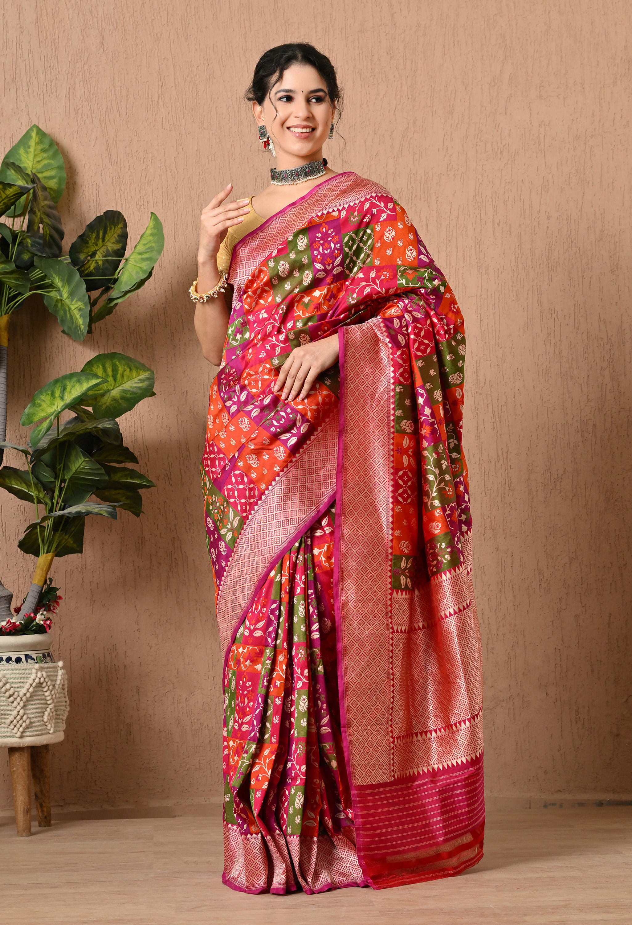 A woman smiling and posing in a traditional Rangkat Pure Katan Silk Saree from The Weavers Shop by Raavi. The handwoven saree features a vibrant multi-coloured palette of magenta, orange, and olive green in a diagonal block pattern. It is adorned with intricate gold zari work consisting of delicate floral and leafy motifs throughout the body. The saree is finished with a broad, ornate gold zari border and a rich magenta pallu. This Silk Mark Certified garment is paired with a gold blouse and a silver choker