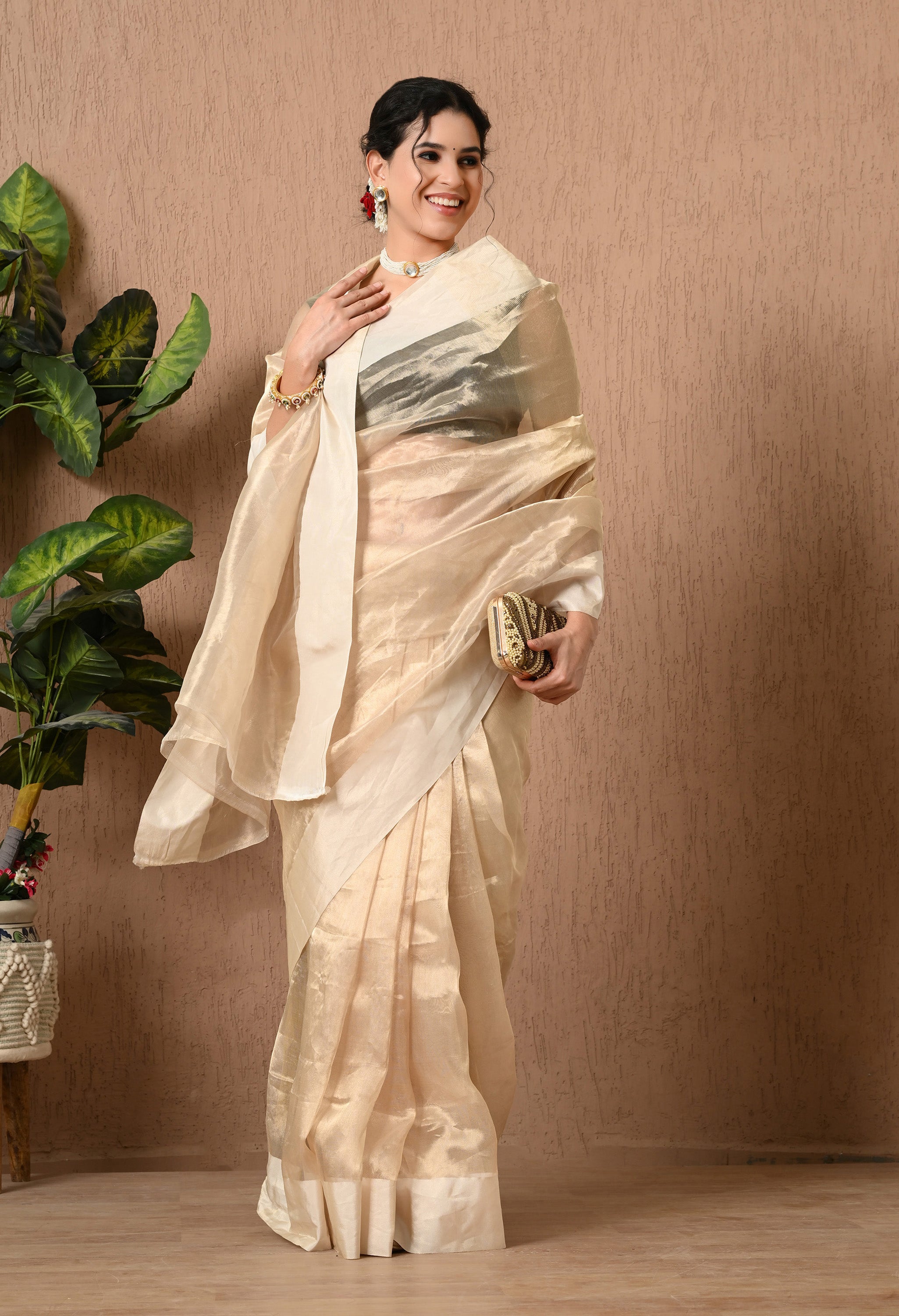 A woman smiles while posing in a shimmering, champagne-gold tissue silk saree from The Weavers Shop by Raavi. The translucent pallu is elegantly draped over her left shoulder, highlighting the fabric's delicate sheen against a dark sleeveless blouse. She is styled with a pearl choker and an embellished clutch, standing against a neutral textured wall.