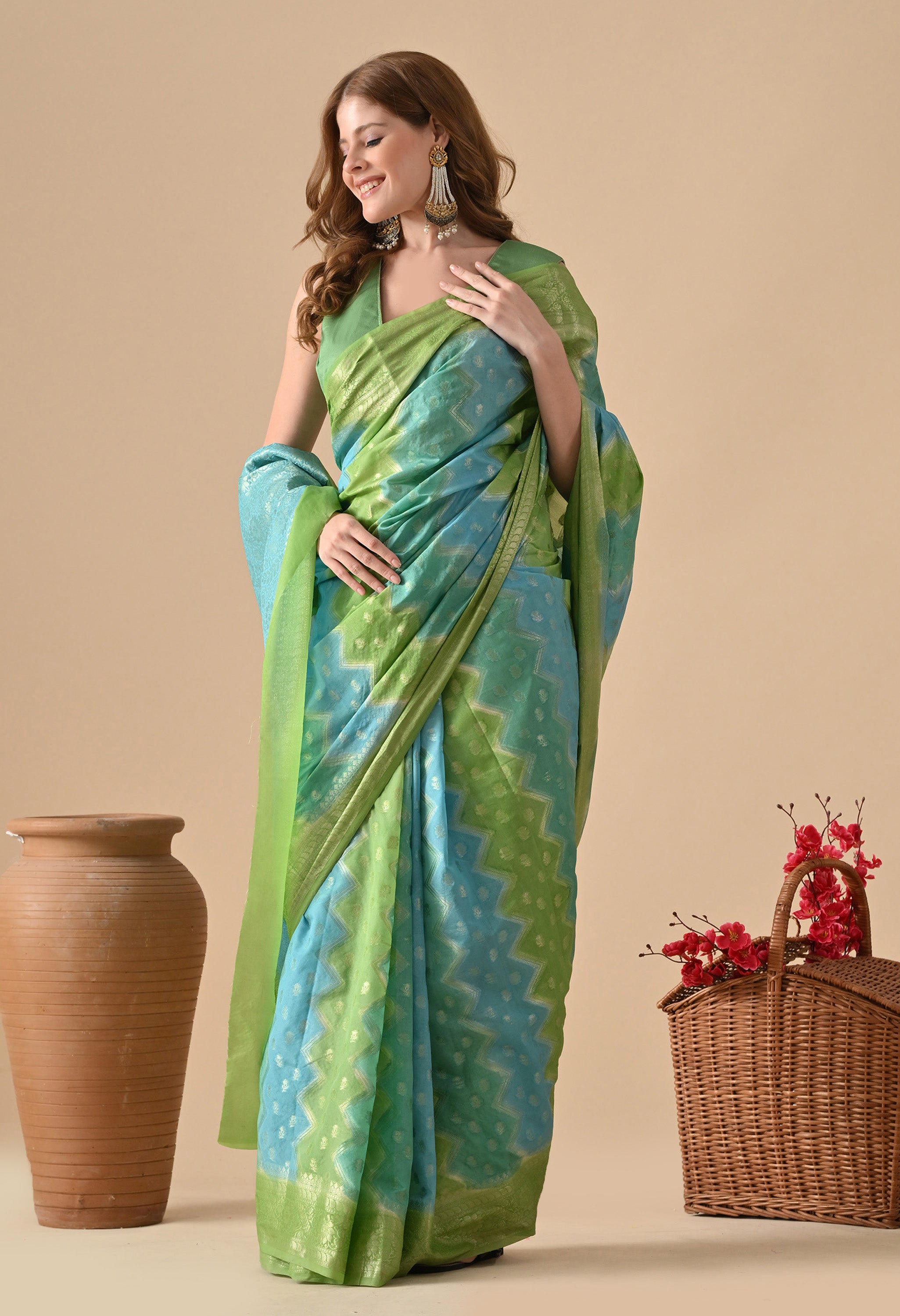A woman stands in a three-quarter profile, smiling and looking down, while wearing a seafoam green and sky blue Banarasi Rangkat saree from The Weavers Shop. The saree features a distinctive diagonal zigzag pattern and is embellished with delicate gold zari buttas and a shimmering gold border. She is styled with long, ornate statement earrings and a matching green sleeveless blouse. The background is a neutral beige, accented by a rustic clay vase and a basket of pink-red flowers.
