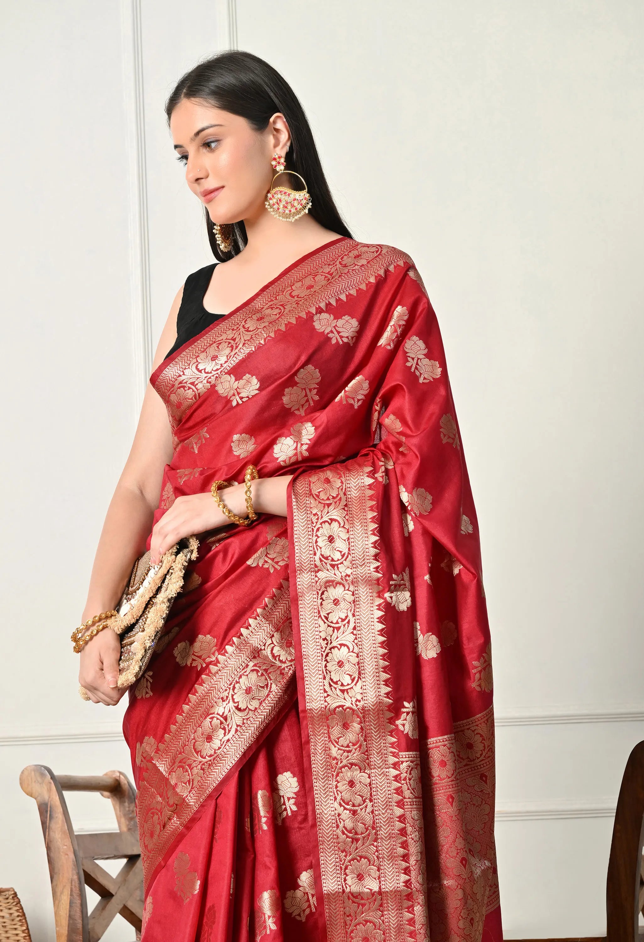 A mid-closeup shot of a woman looking down and to the side, wearing a luxurious crimson red Banarasi silk saree. The saree features intricate golden zari work, including detailed floral butta patterns across the fabric and a broad, ornate gold border with traditional motifs. She has styled the saree with a sleeveless black blouse, large statement earrings, and gold bangles, while holding clutch. The background is a clean, white 